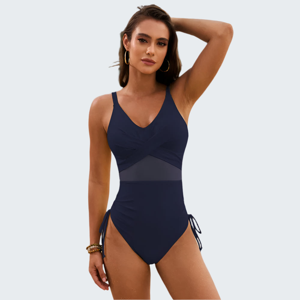 A woman standing on a beach wearing a navy blue one-piece swimsuit with a v-neck and drawstrings on the sides.
