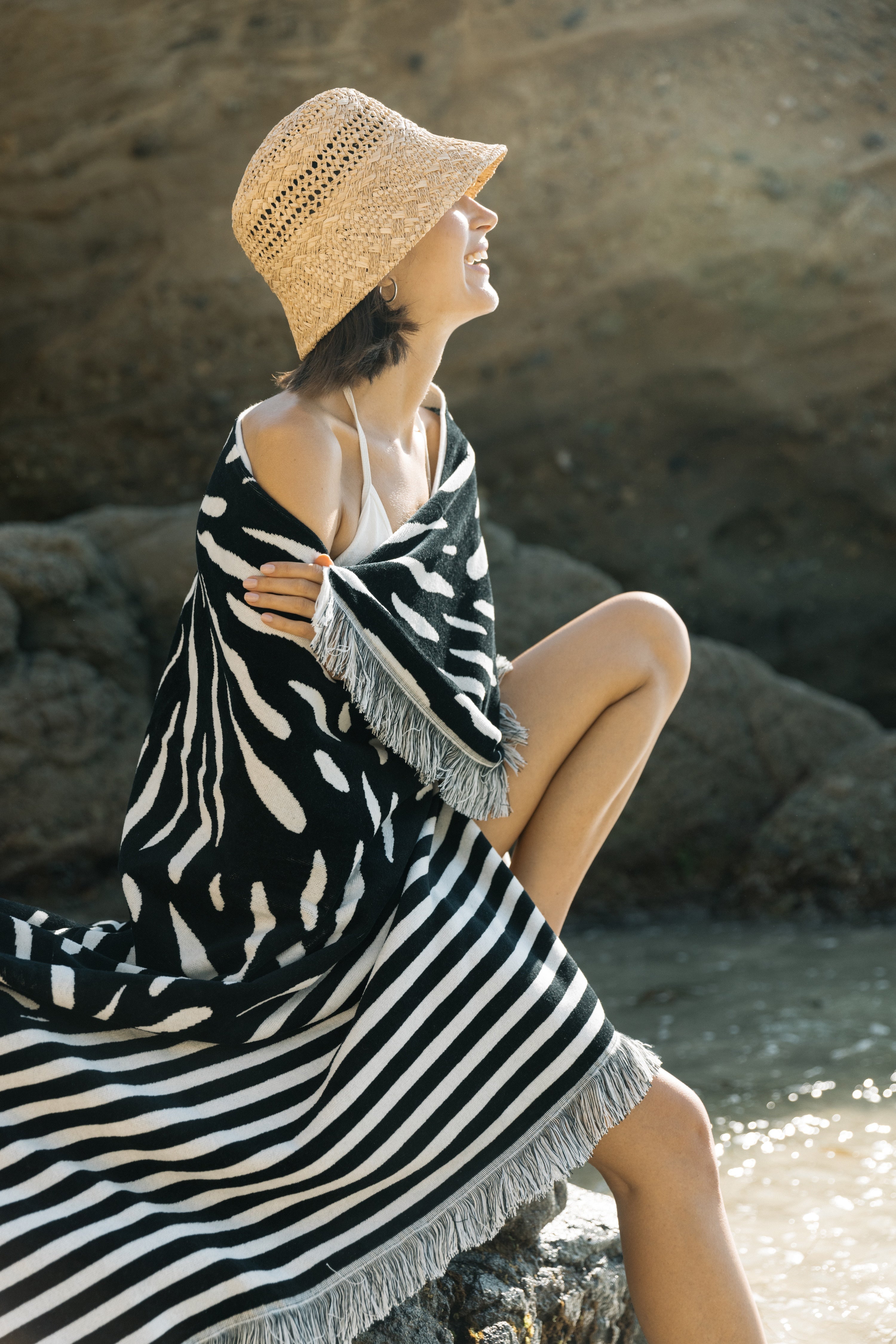 Woman sitting on rocks by the water wearing a black and white striped towel and straw hat.