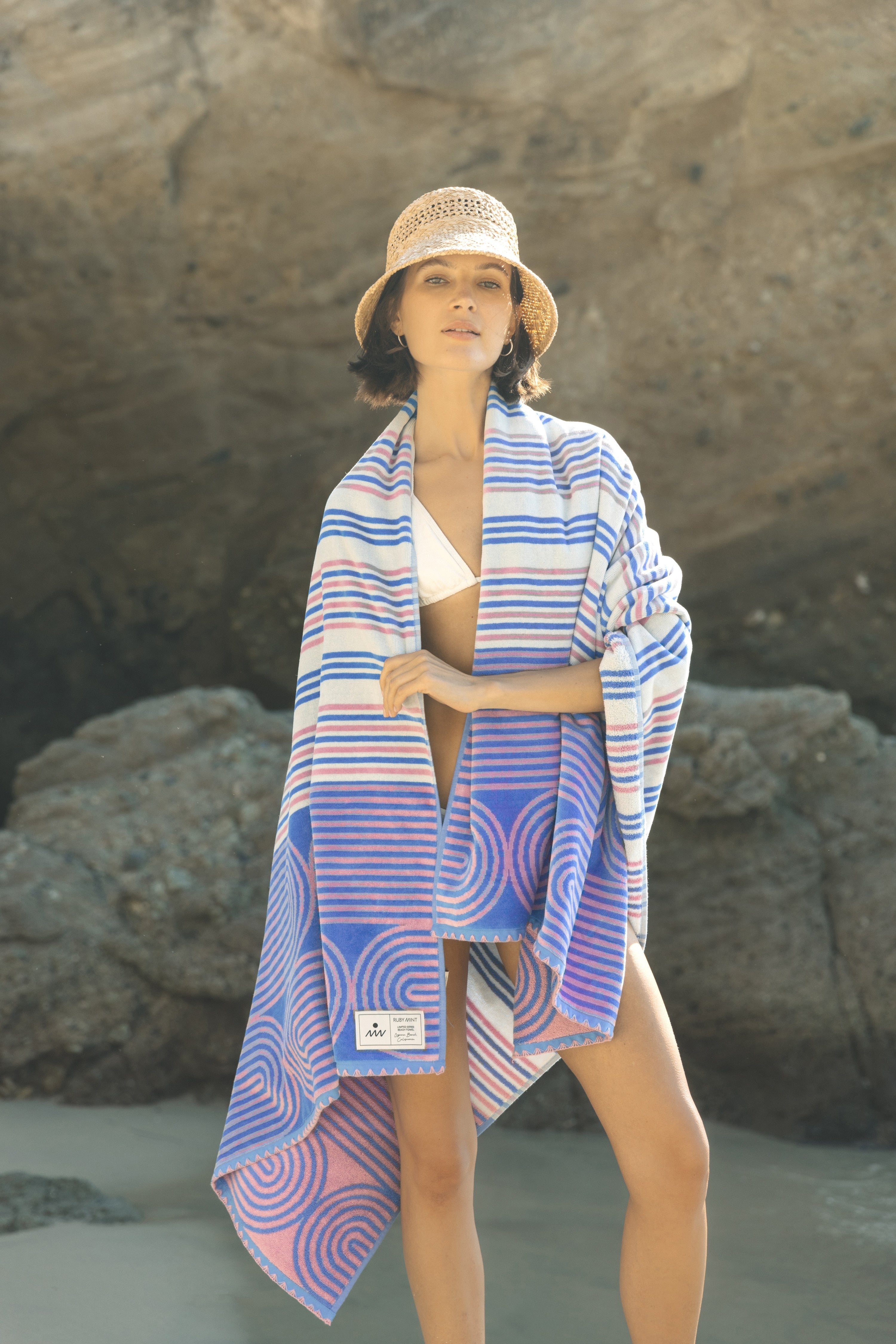 Woman on a beach wearing a blue and pink patterned towel draped over her shoulders.