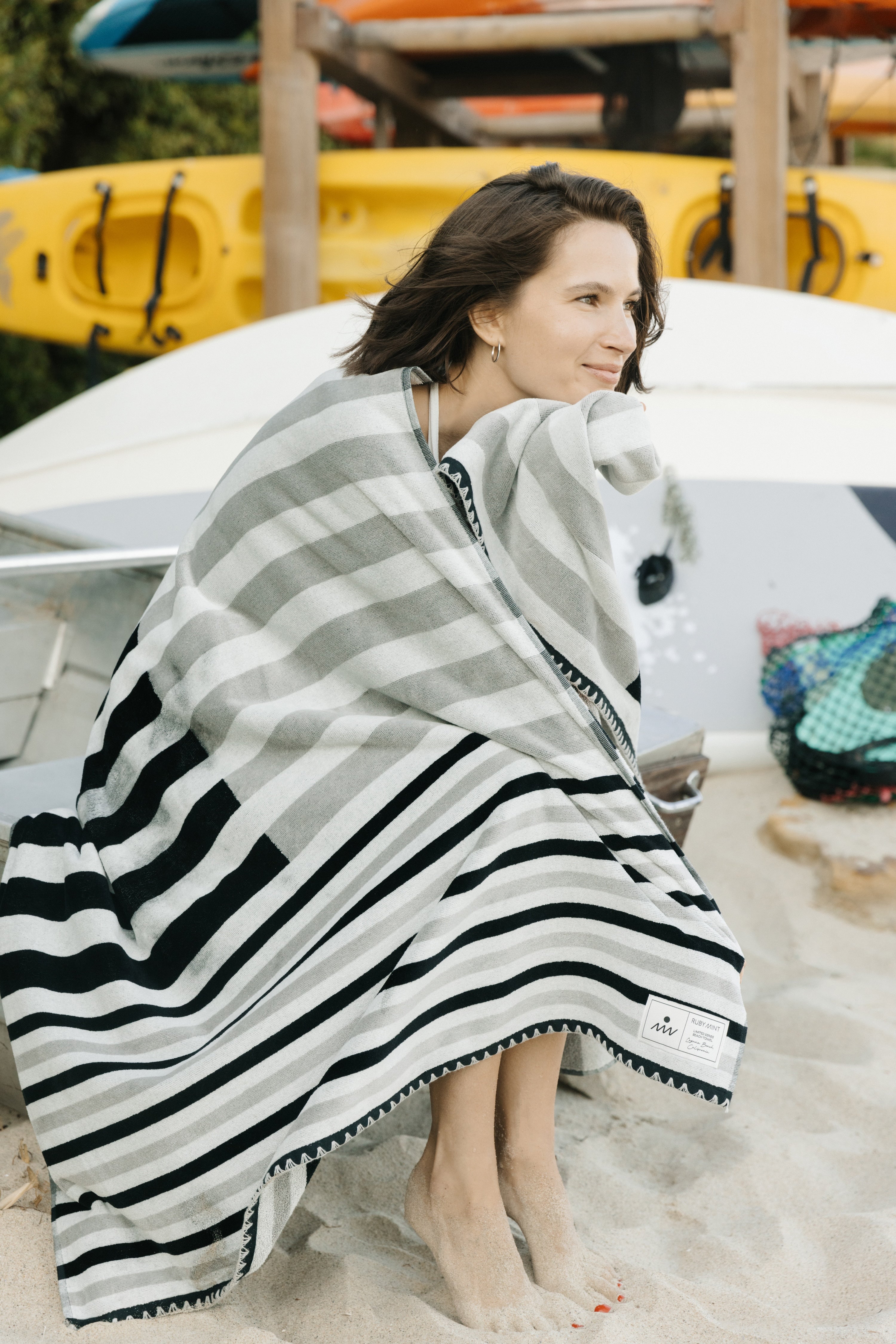 Woman wrapped in a striped towel on a sandy beach with kayaks in the background