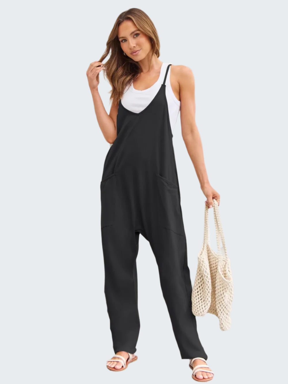 A woman standing on a patio wearing a black sleeveless jumpsuit with a white tank top underneath, holding a wicker handbag and wearing white sandals.