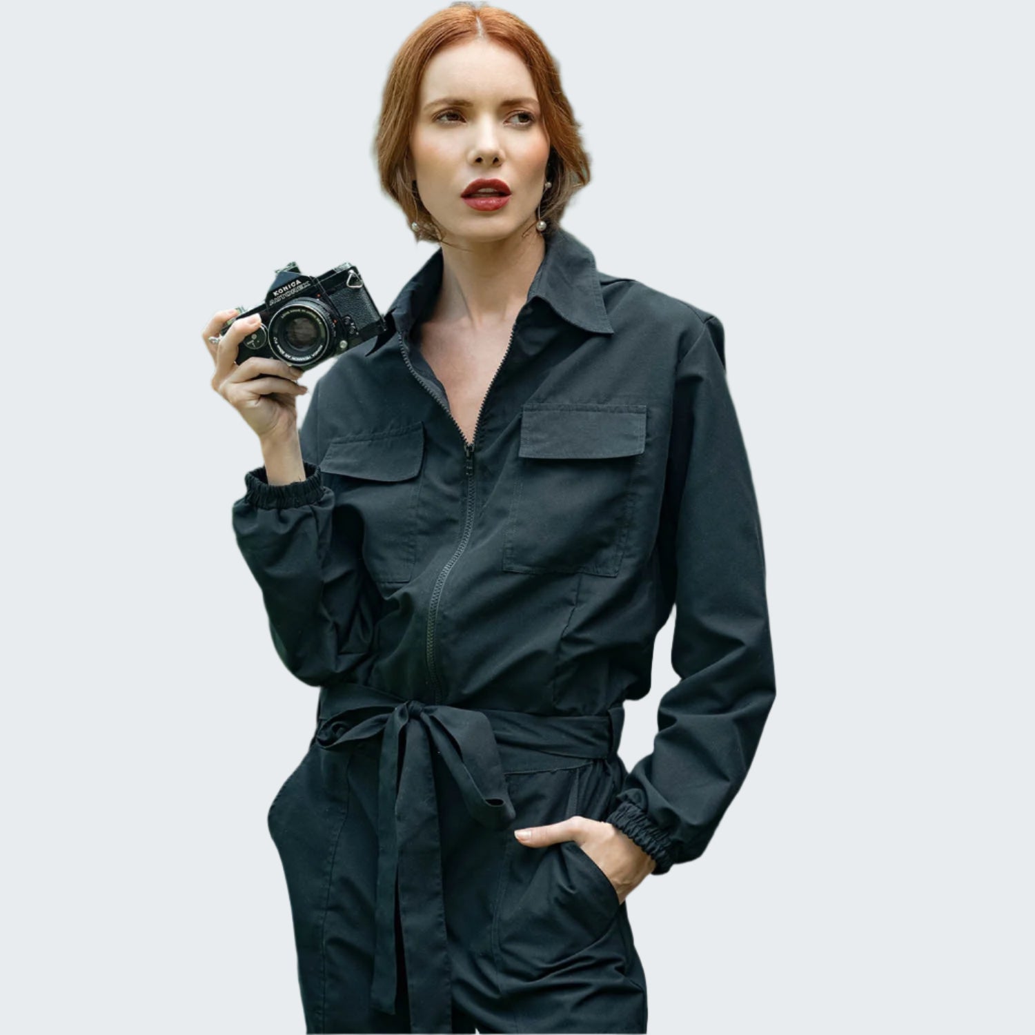 Woman in a black outfit holding a camera in an outdoor setting with greenery.