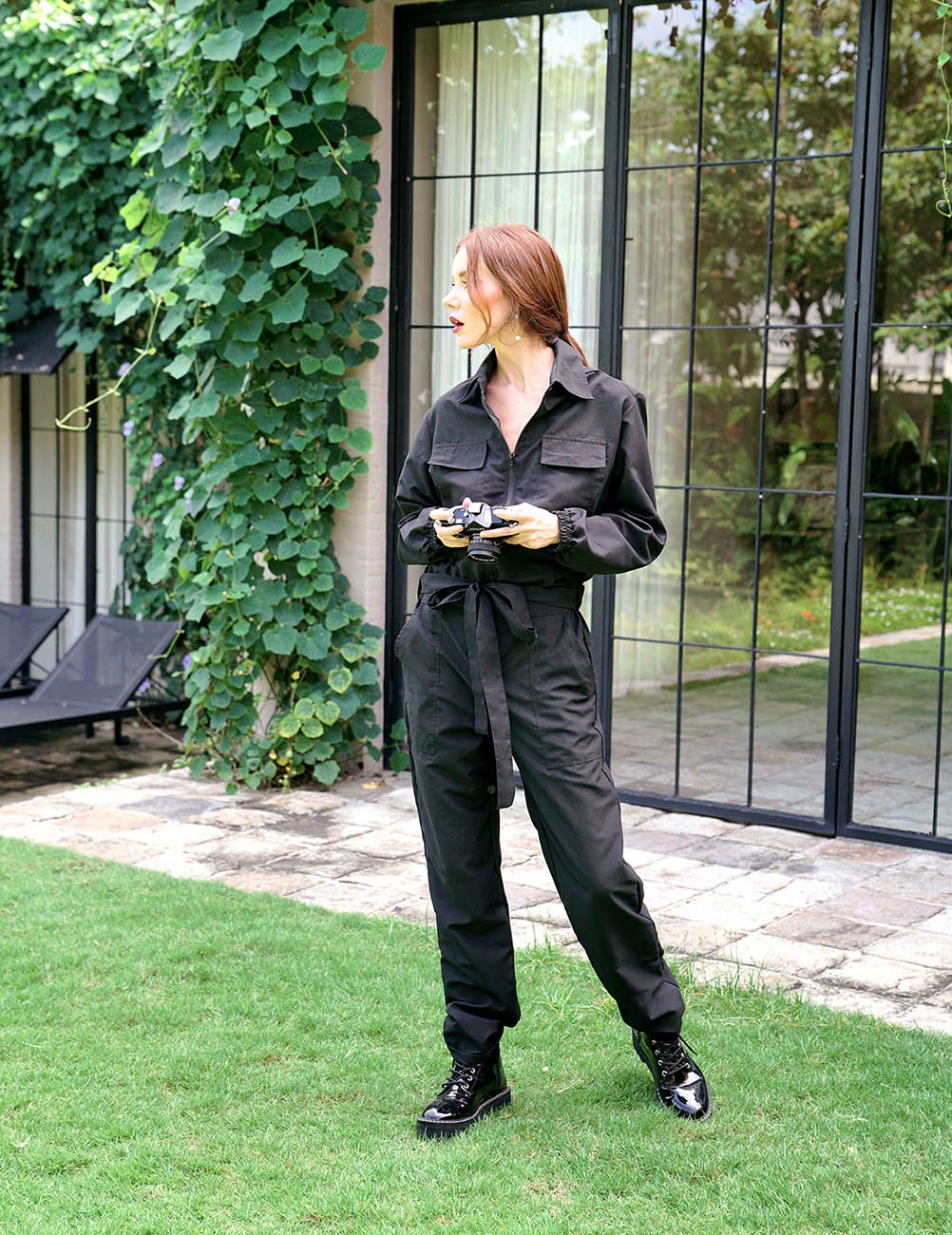 Woman in a black jumpsuit standing outdoors near a building with greenery.