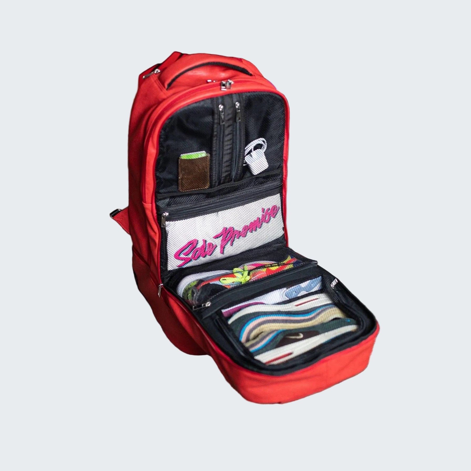 Red backpack with open compartments on a wooden floor, with shoes and boxes in the background.