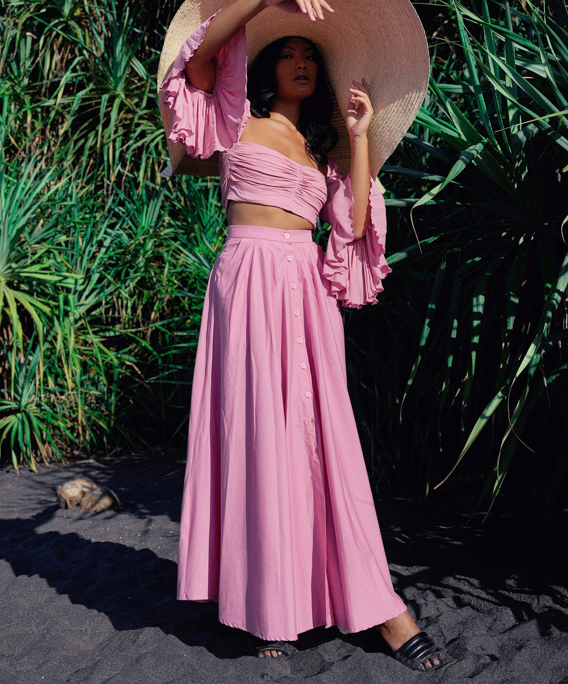 Woman in a pink outfit with a wide-brimmed hat standing on a sandy beach with greenery in the background.