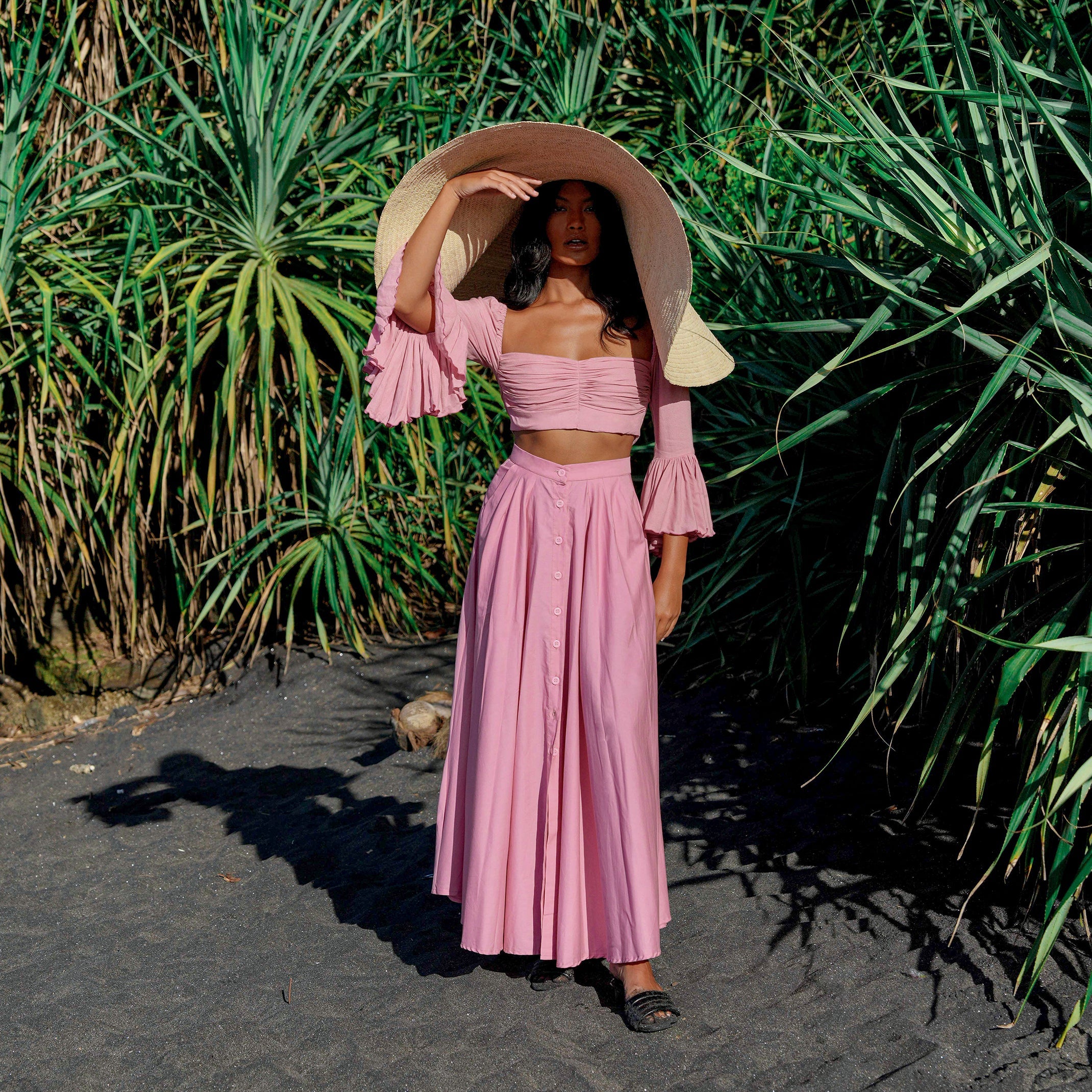 Woman in a pink outfit with a wide-brimmed hat standing in front of green plants.