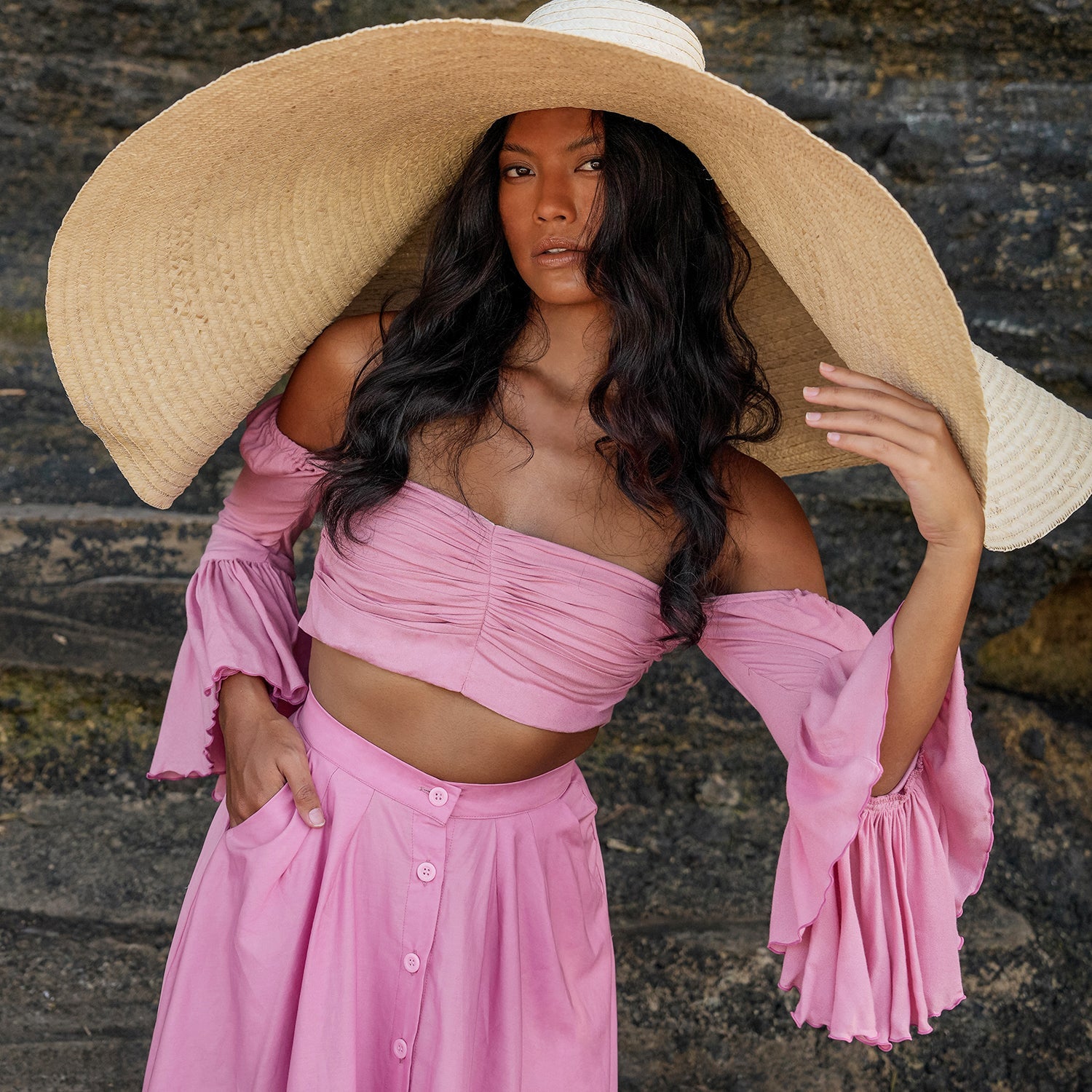 Woman wearing a pink outfit and large straw hat against a stone wall.