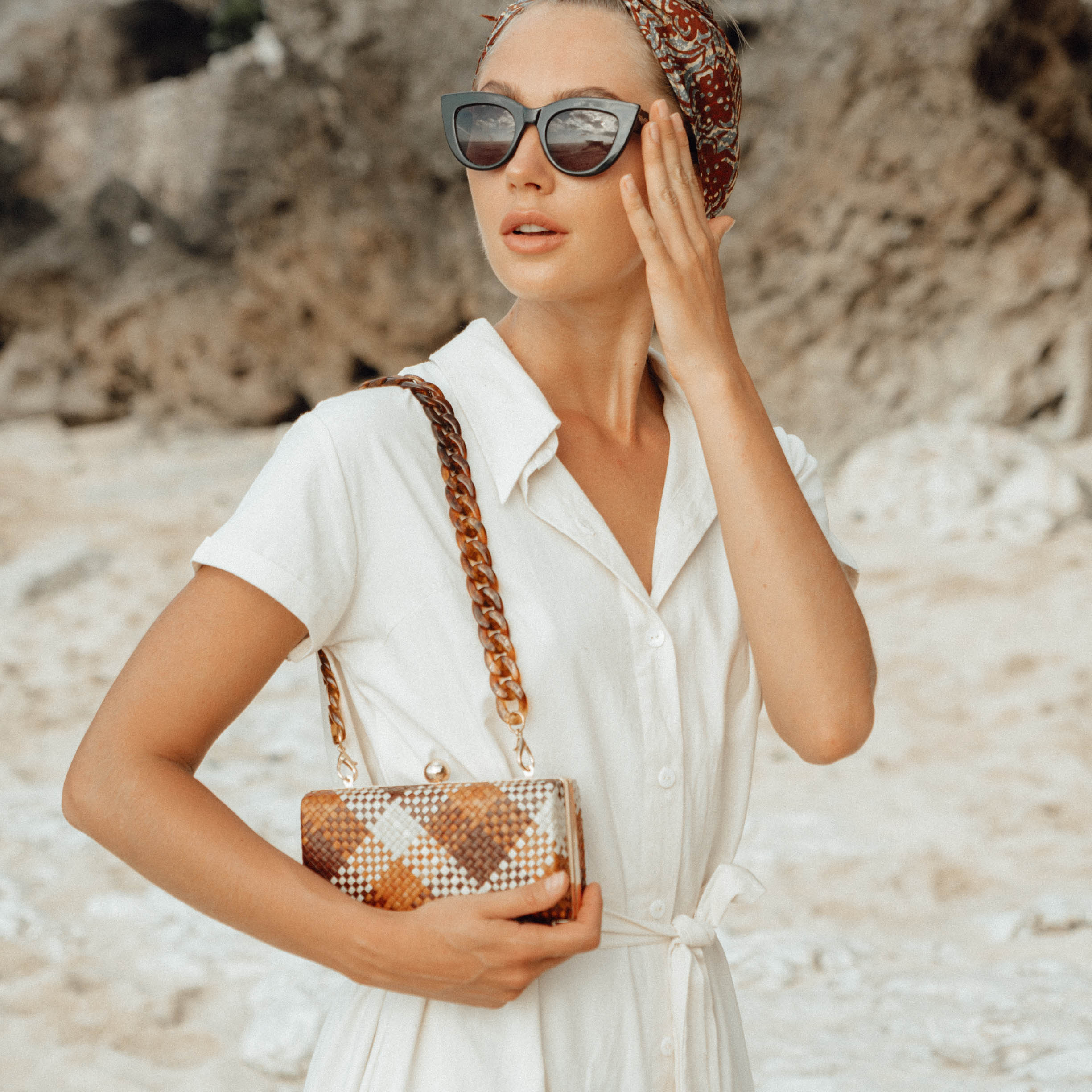 Woman in a white dress holding a patterned handbag on a beach.
