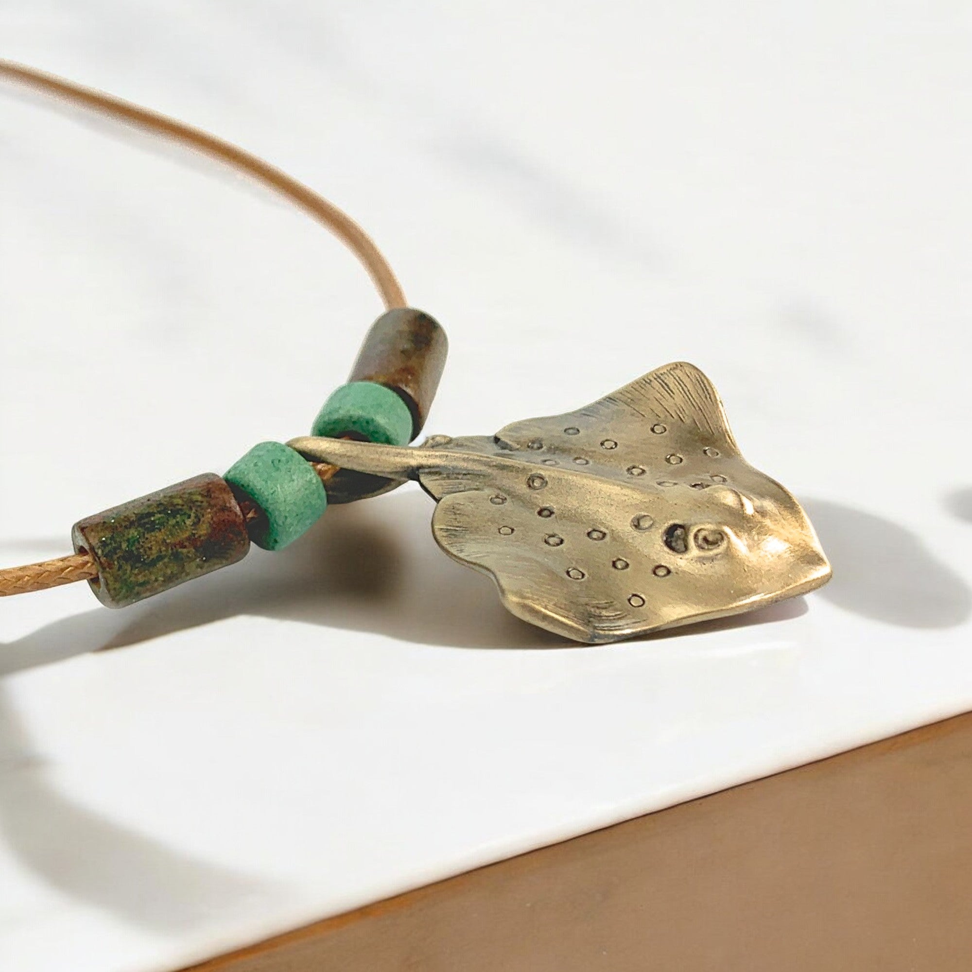 Brass stingray pendant on a cord with green beads against a white background