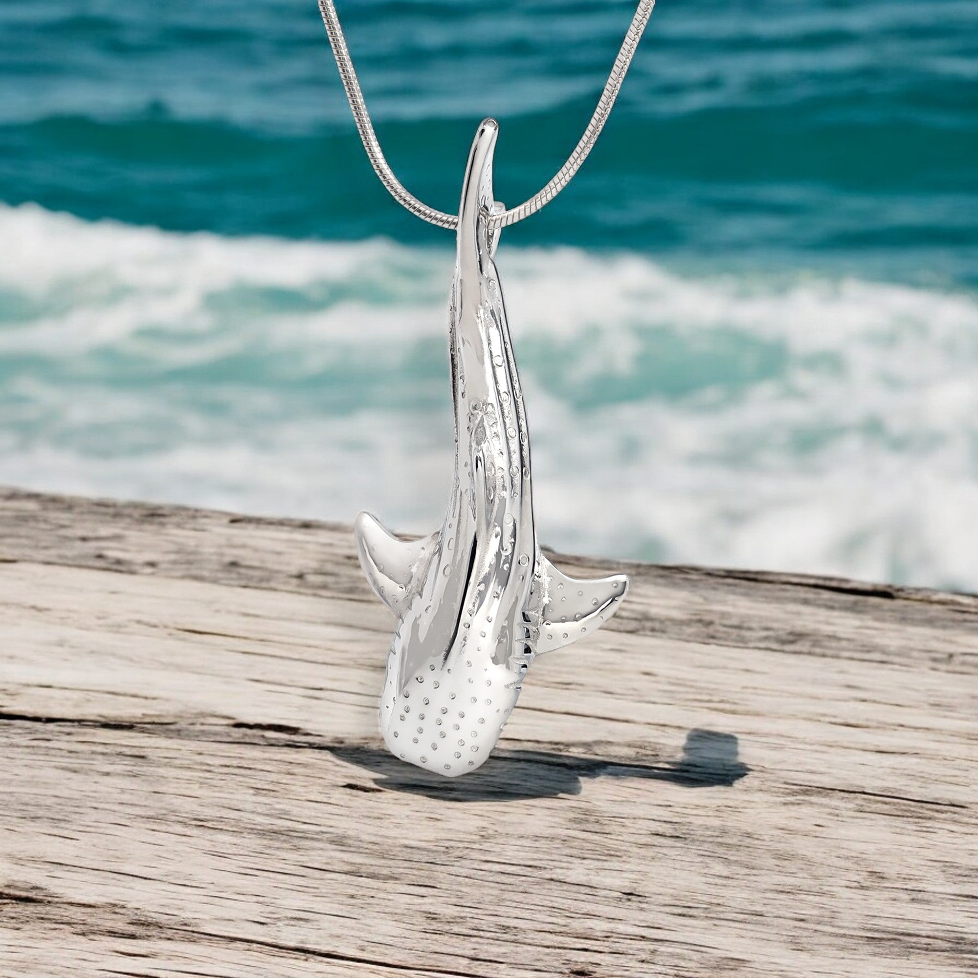Silver shark pendant on a wooden surface with ocean in the background