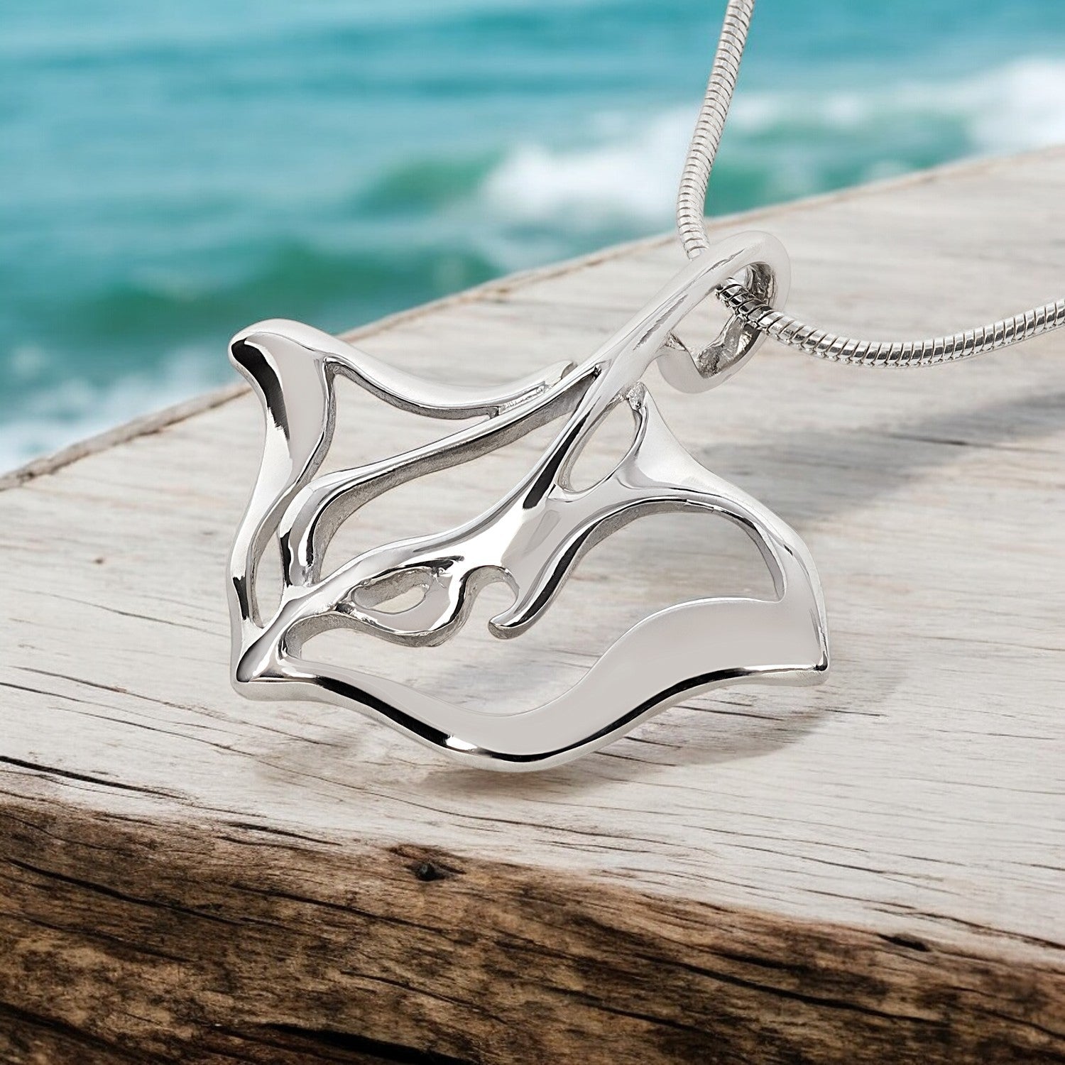 Silver pendant shaped like a stingray on a wooden surface with ocean in the background