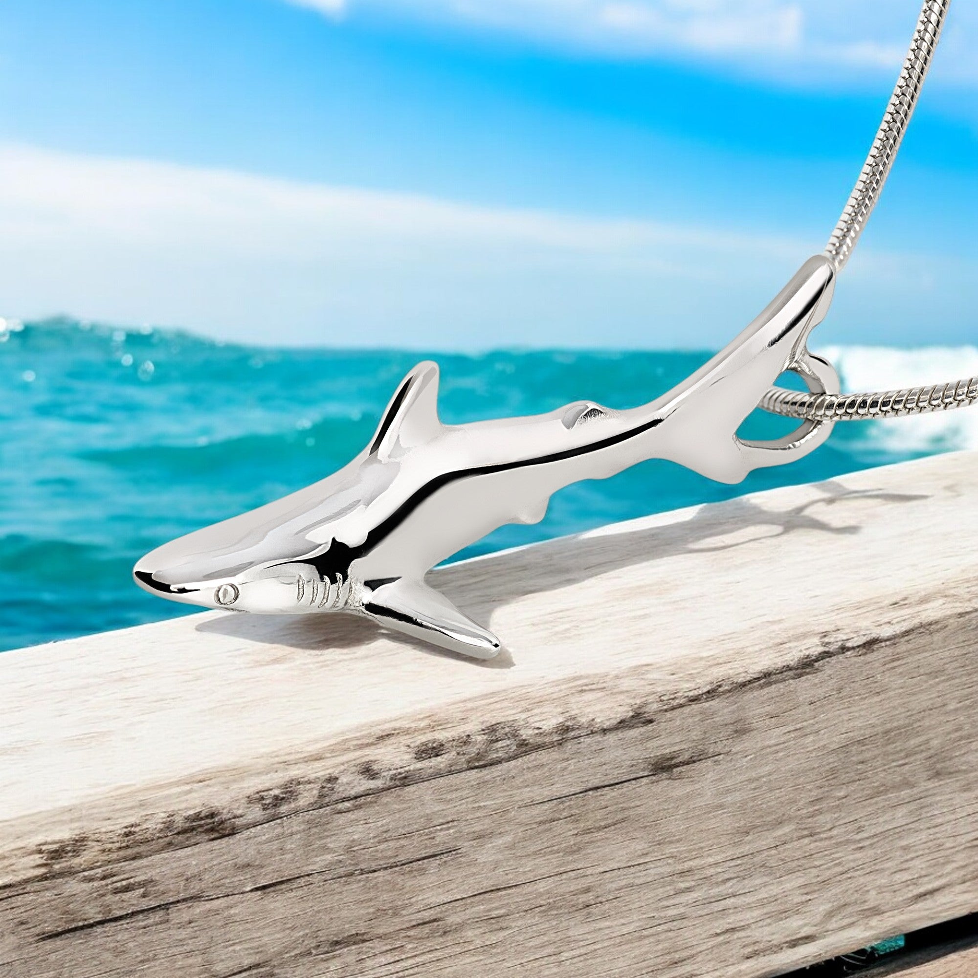Silver shark pendant on a chain against a beach background with ocean and sky.