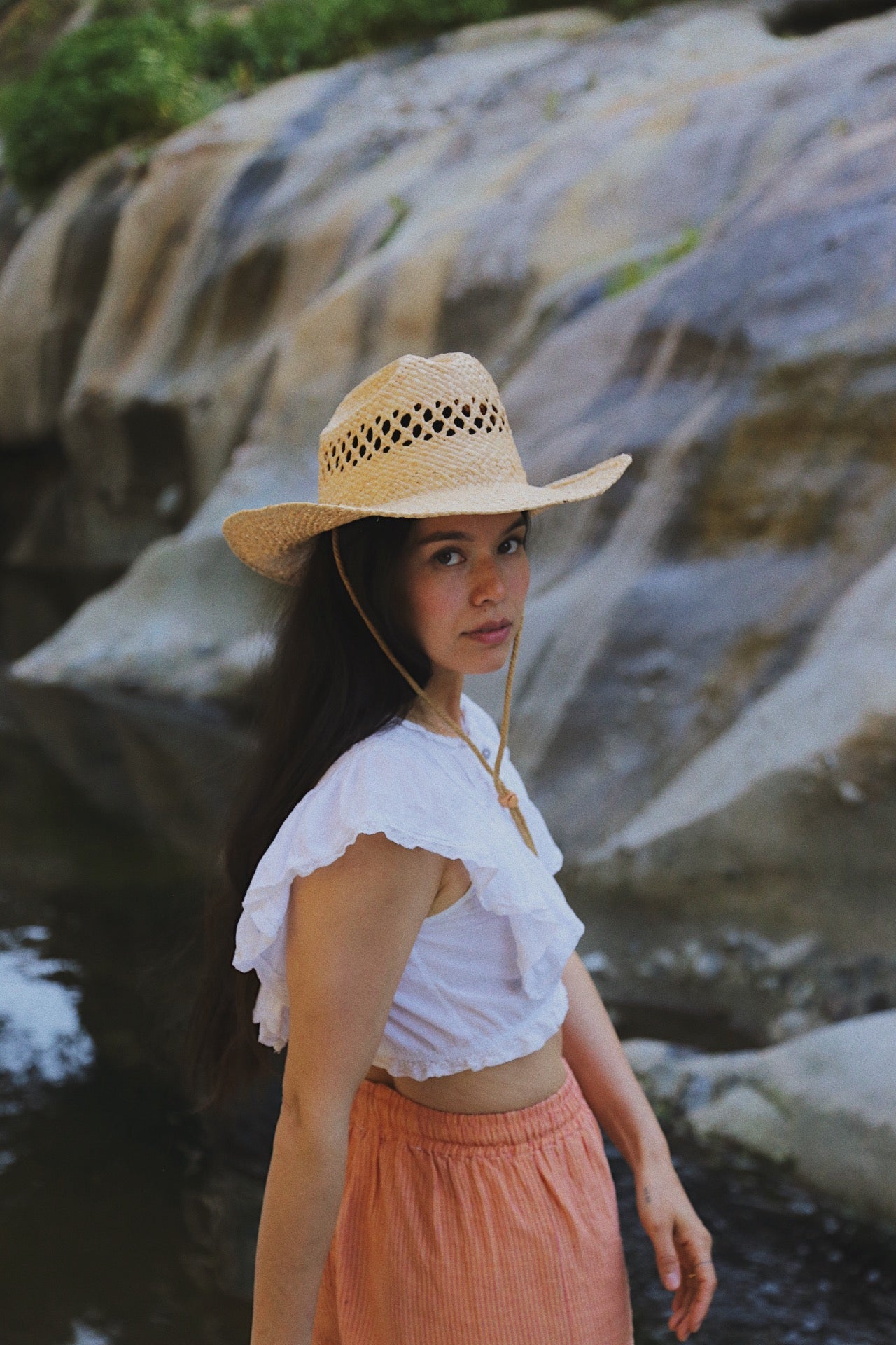 Woman wearing a straw hat and white top with pink pants standing in front of rocky terrain.