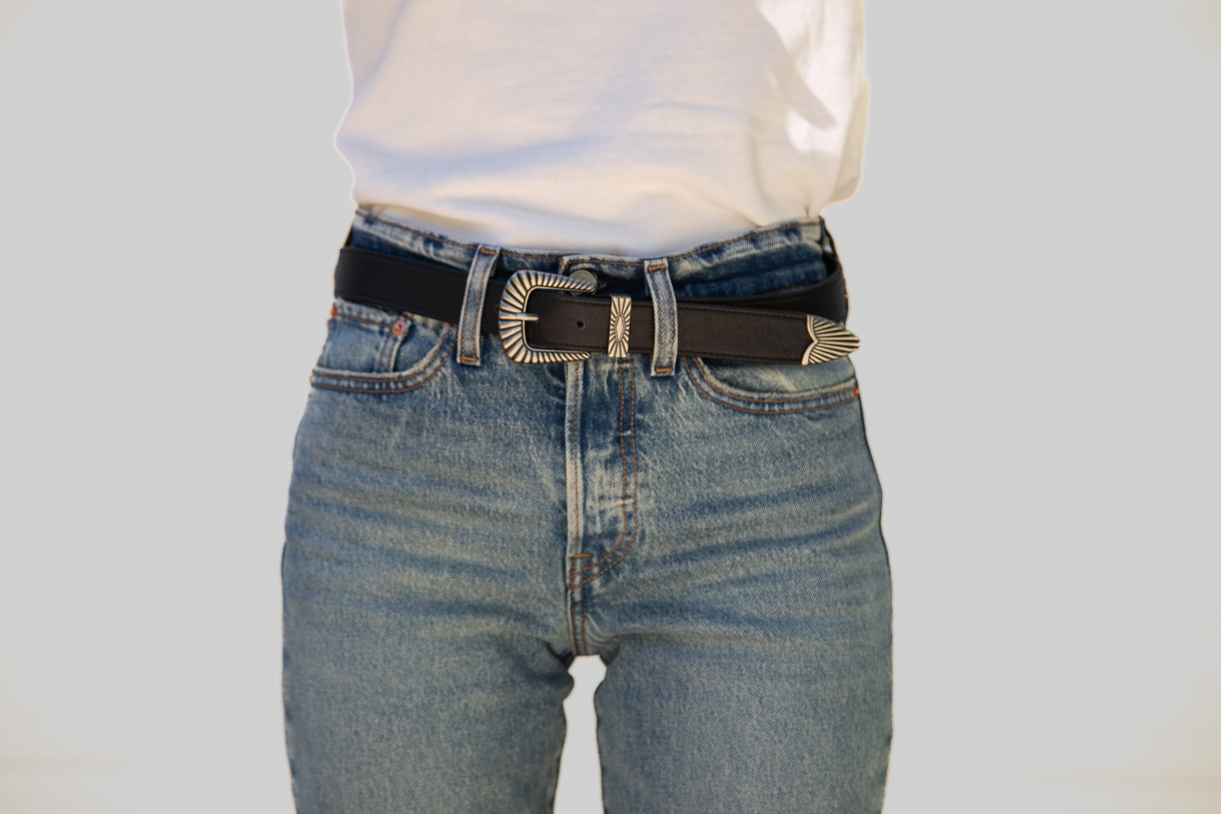 Person wearing blue jeans with a black belt and silver buckle on a light gray background