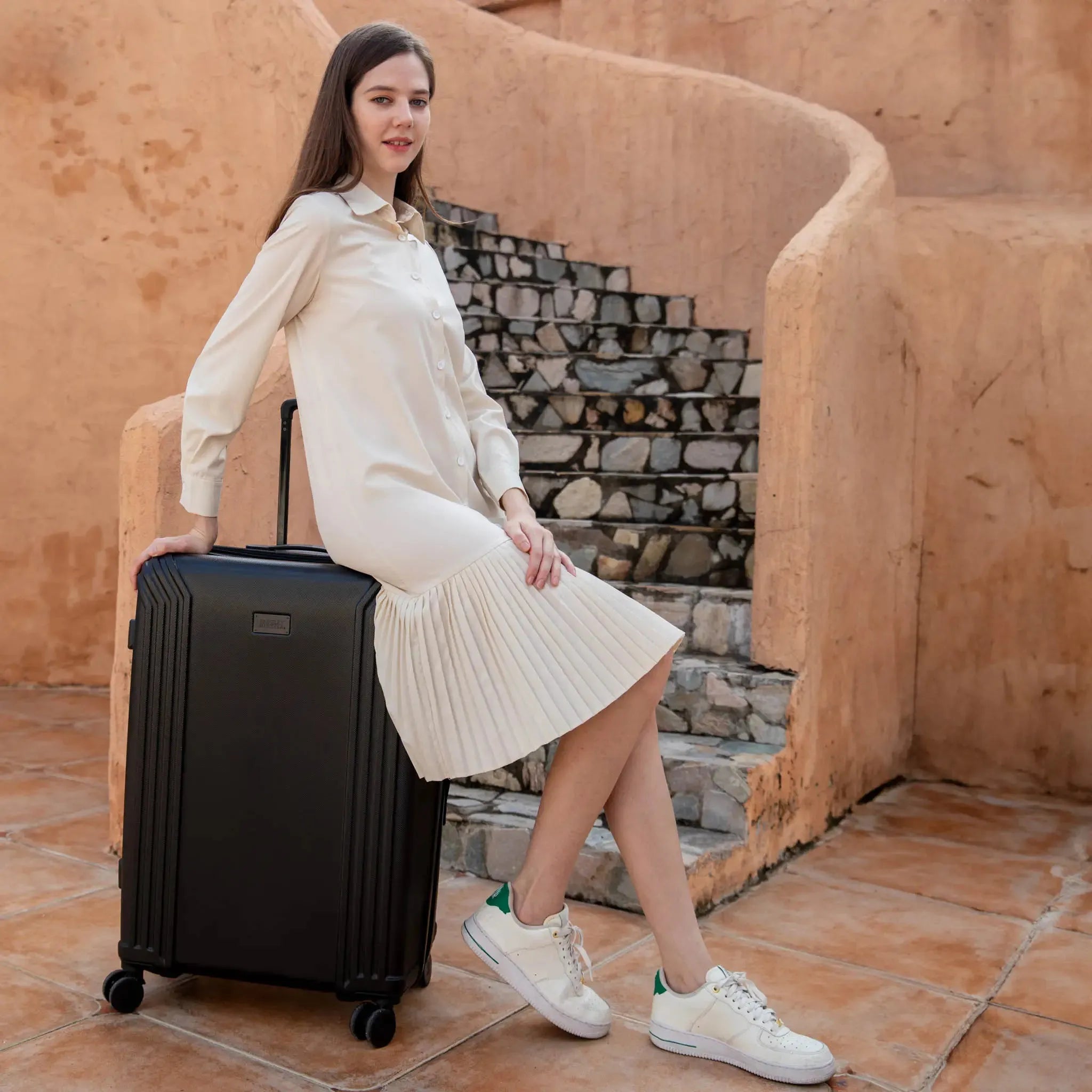 Woman in a white dress sitting on a black suitcase in front of a stone wall.