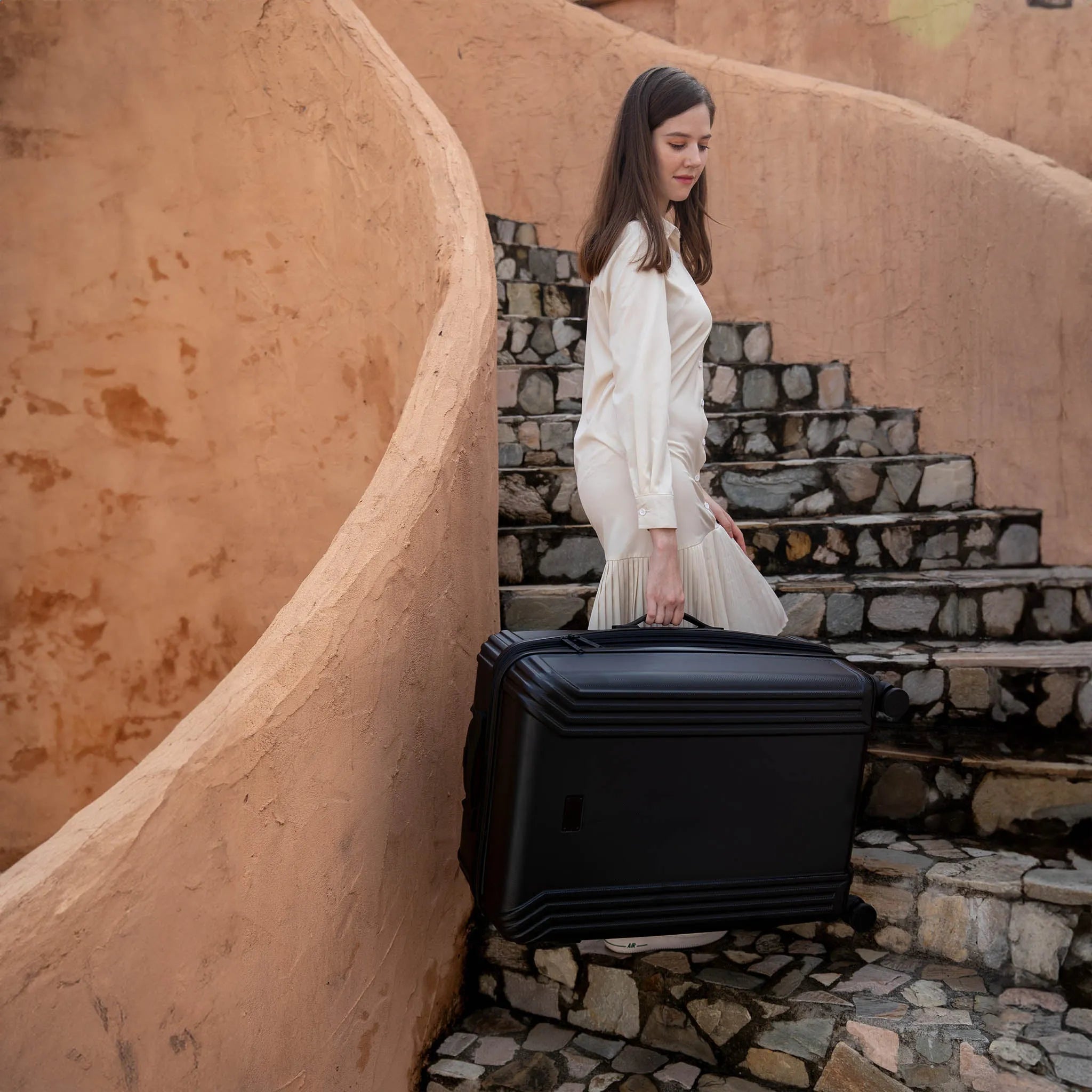 Woman in a white dress holding a black suitcase on stone steps.