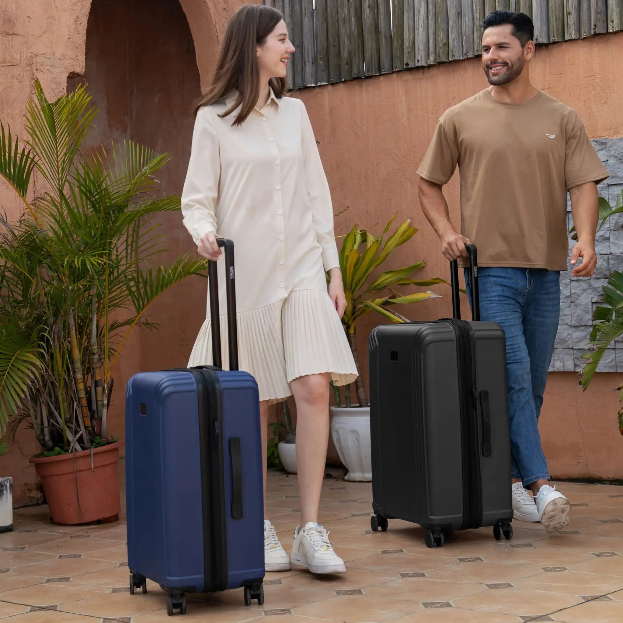 Man and woman walking with suitcases in front of a building with plants.