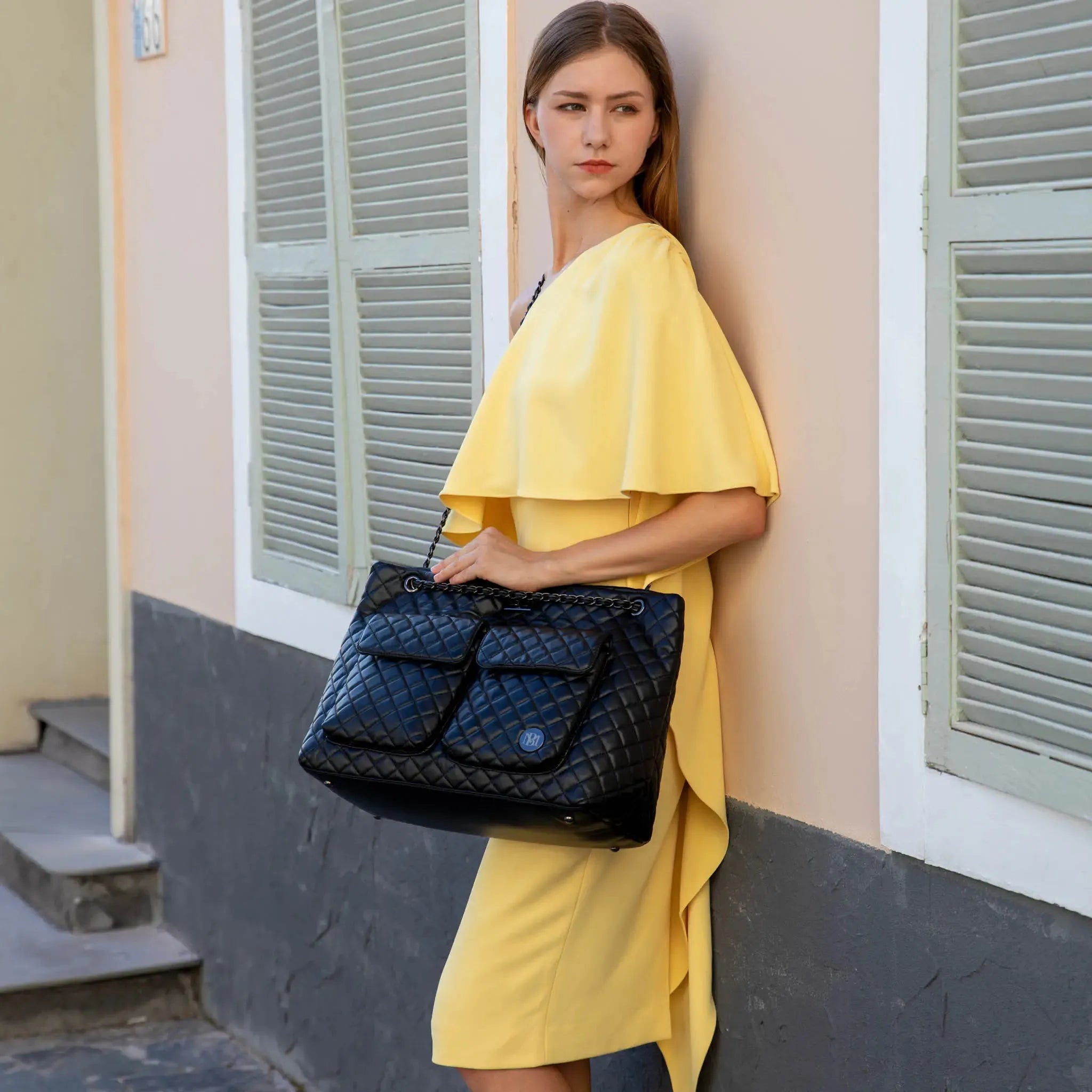 Woman in a yellow dress holding a black quilted handbag against a building background