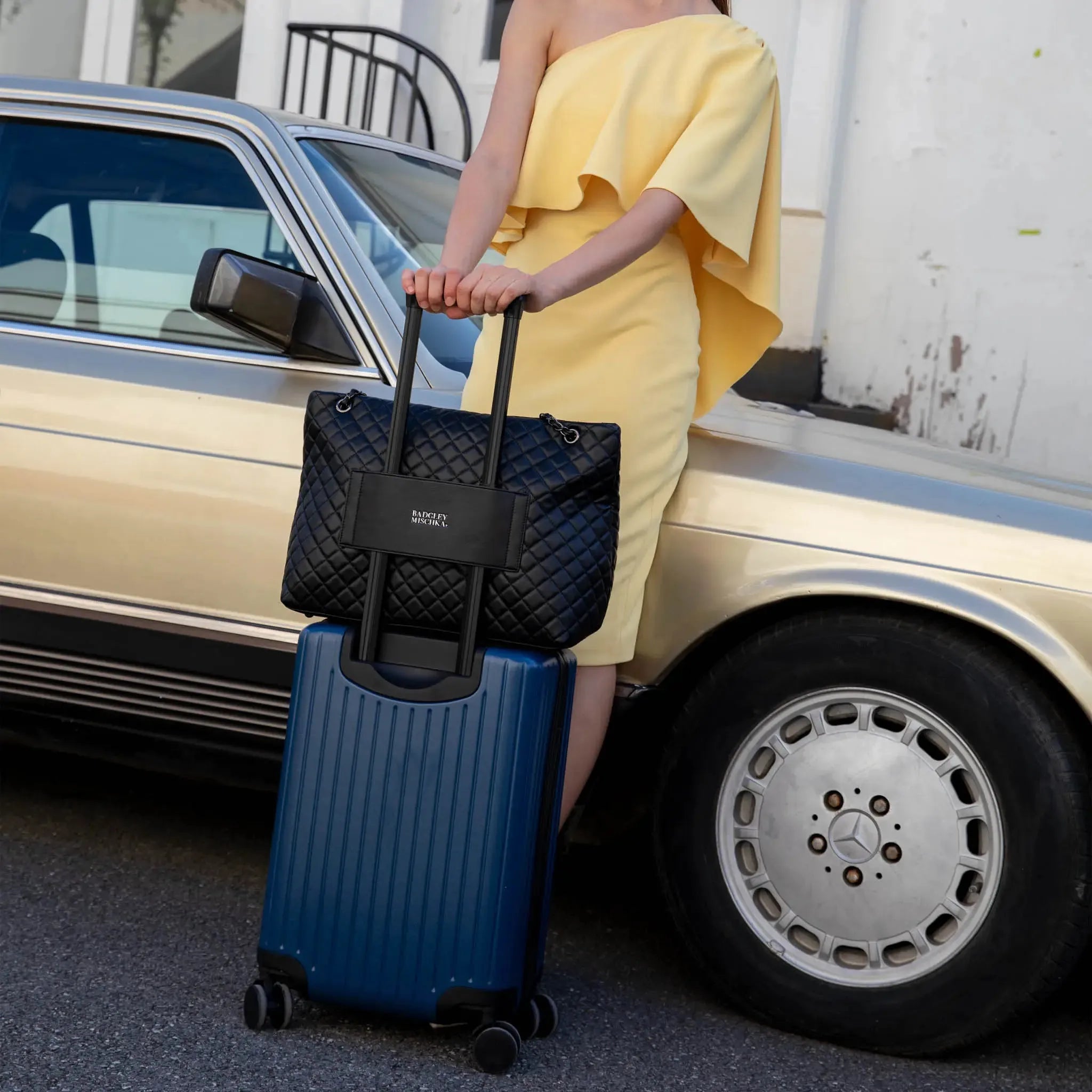 Person holding a black quilted handbag and blue suitcase next to a vintage car.