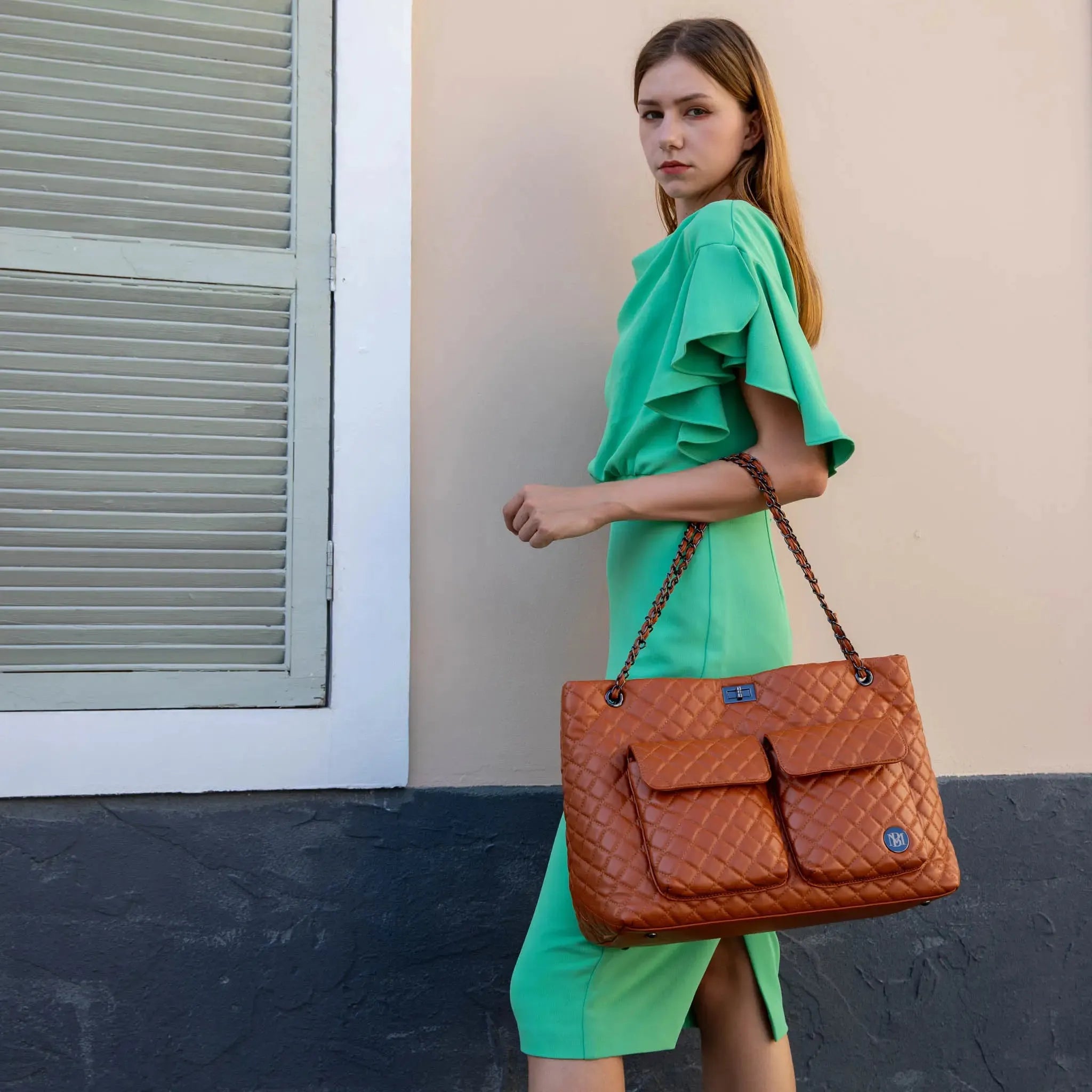 Woman in a green dress holding a brown quilted handbag against a neutral wall.