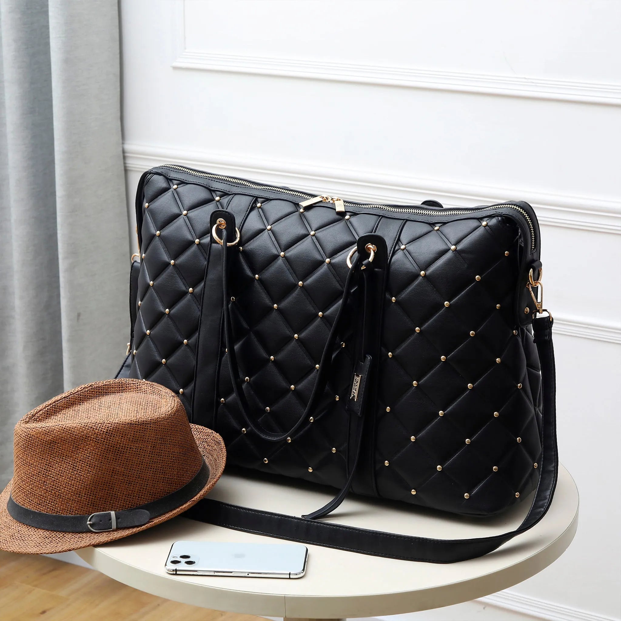 Black quilted handbag with gold studs, brown hat, and smartphone on a white surface.