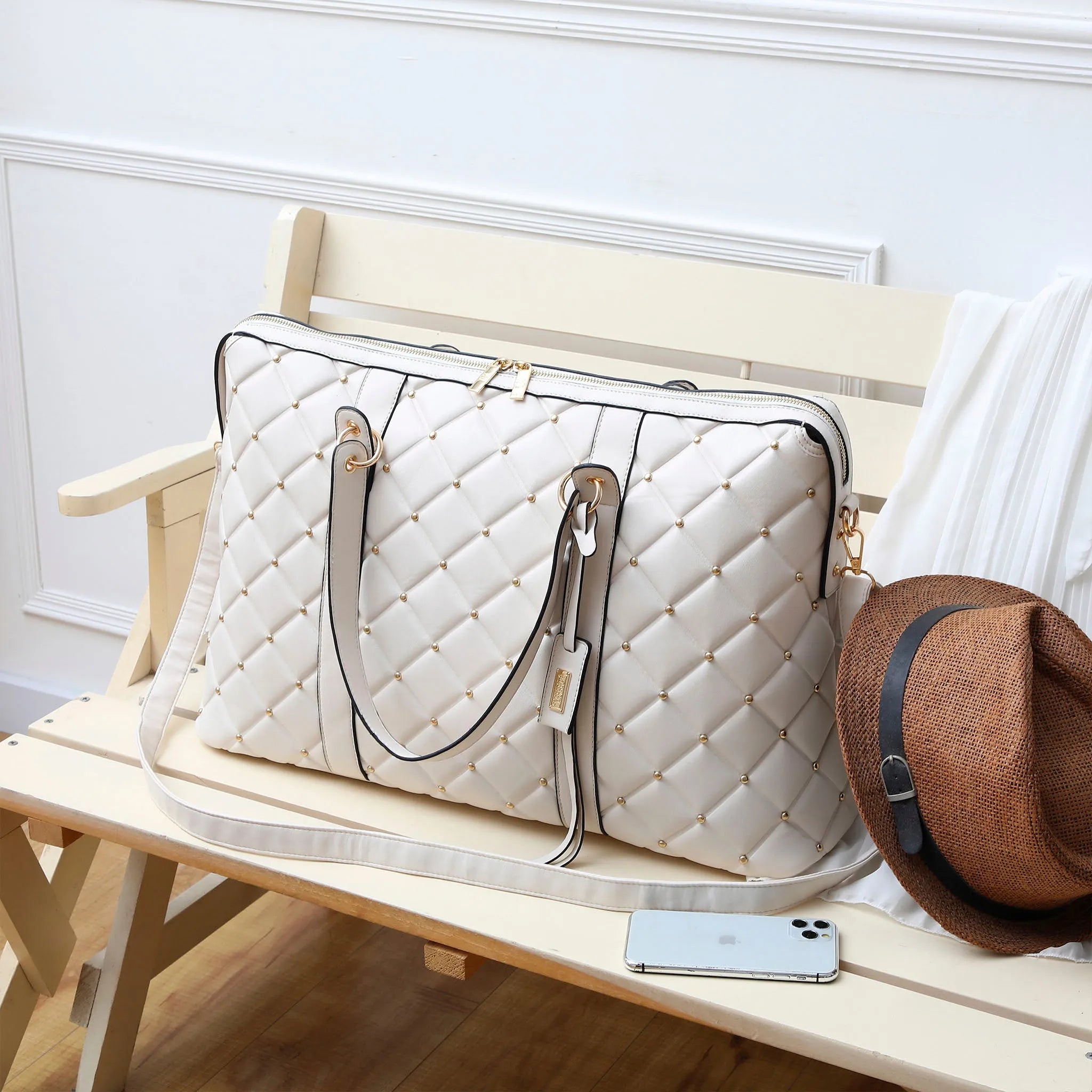 White quilted handbag on a wooden bench with a brown hat and phone beside it.