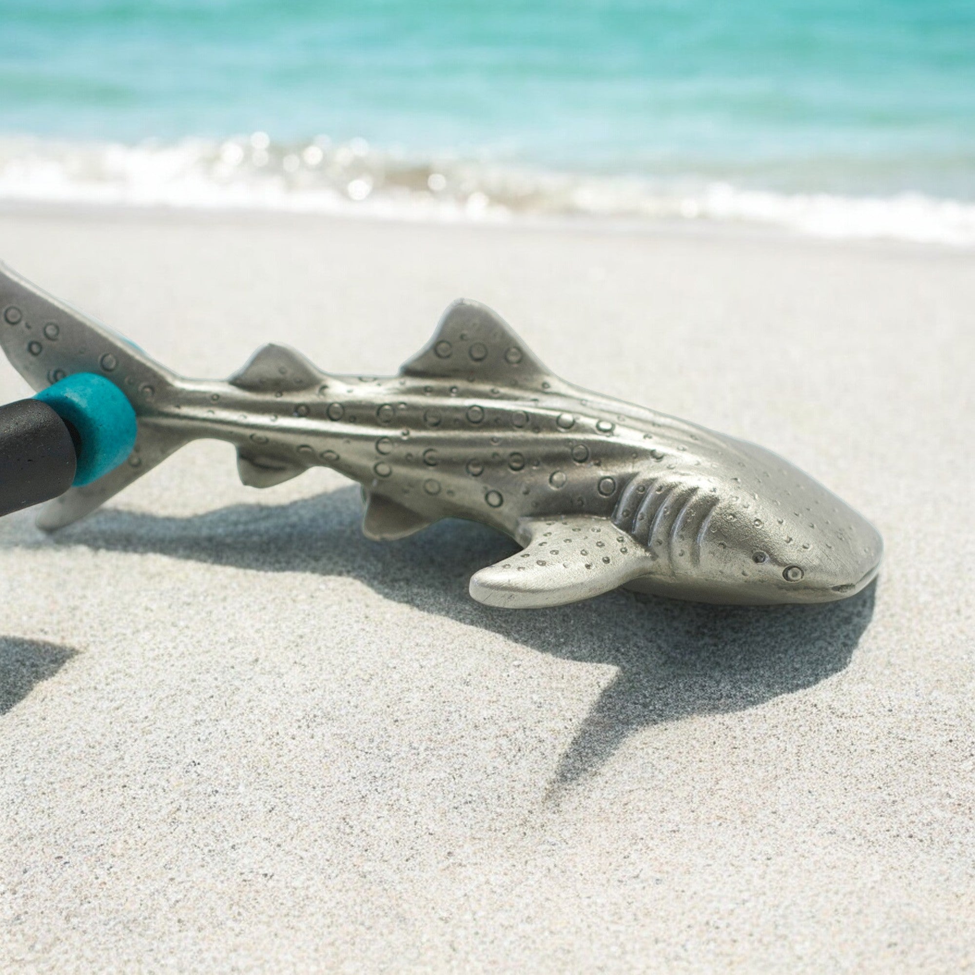 Metallic shark-shaped tool on a sandy beach with water in the background