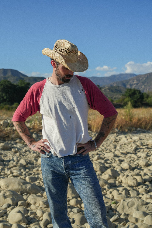 Man wearing a straw hat, red shirt, and blue jeans standing on rocky terrain with mountains in the background.