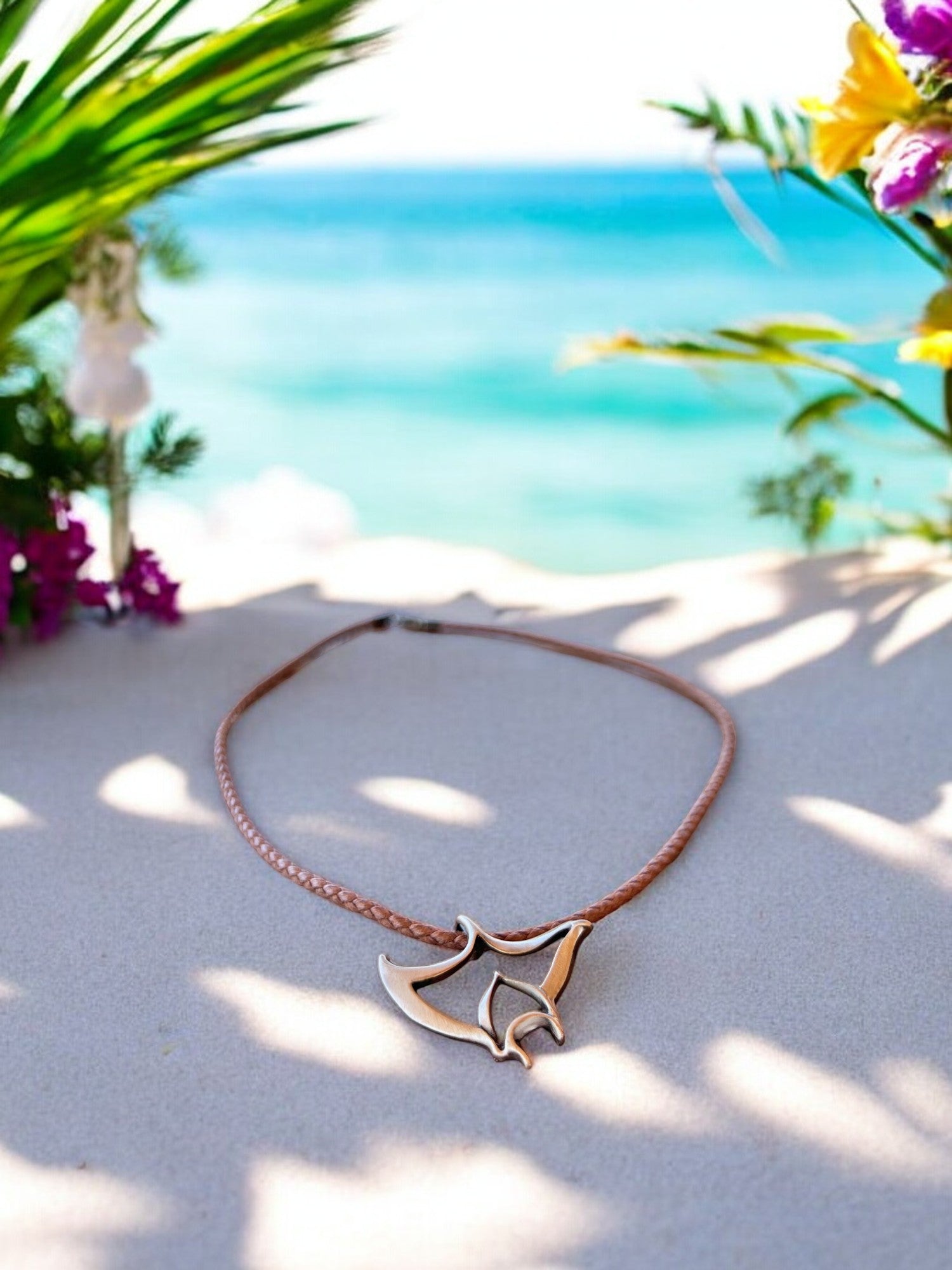 Necklace with a leaf-shaped pendant on a sandy beach with flowers and ocean in the background