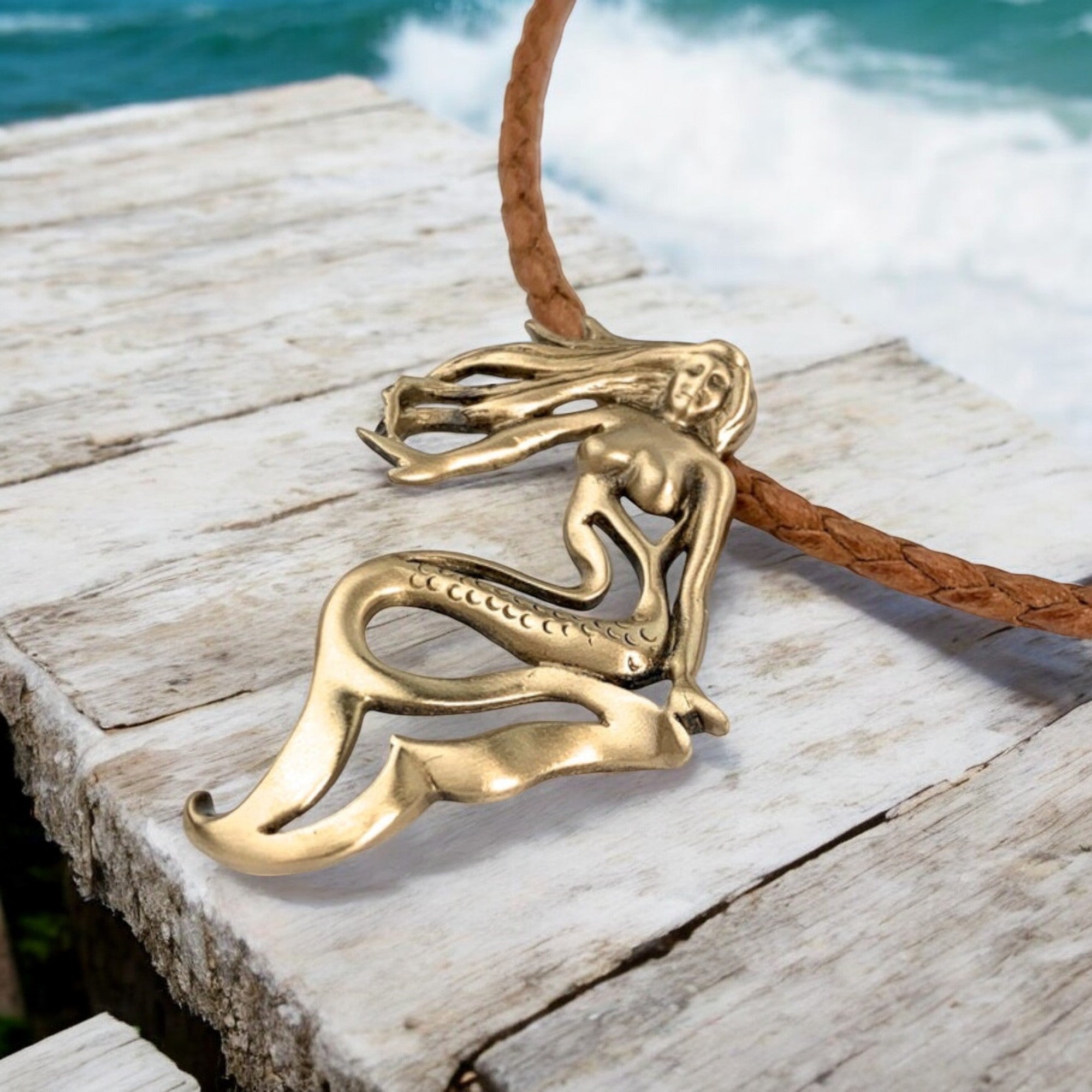 Brass mermaid pendant on a leather cord against a wooden dock background.