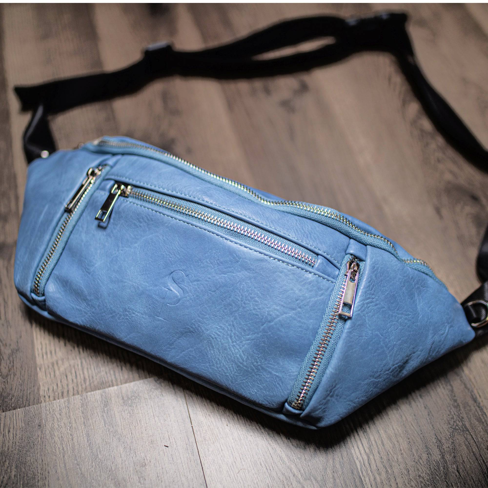 Blue leather waist bag with multiple zippers on a wooden floor