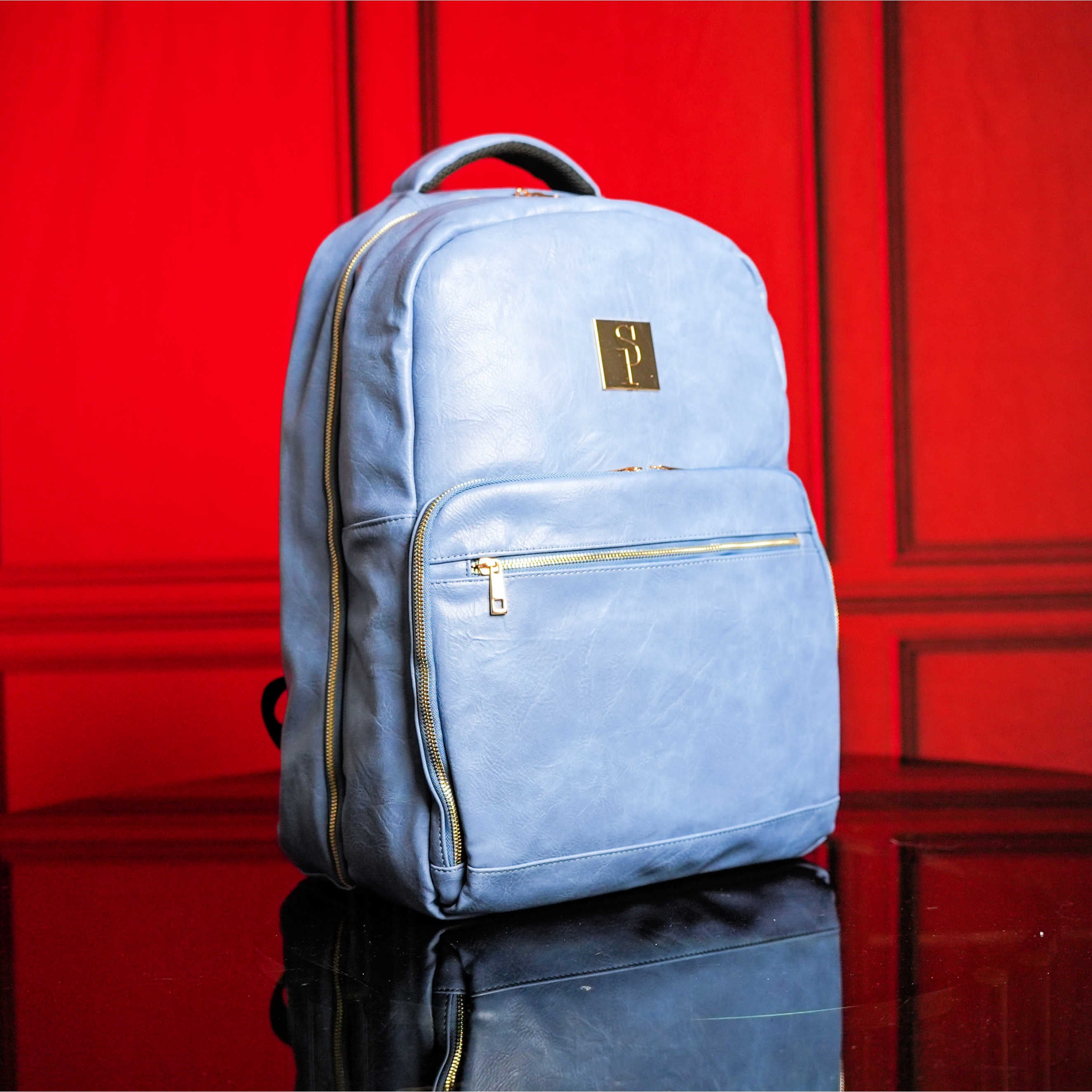 Light blue backpack with gold accents on a reflective surface with a red background