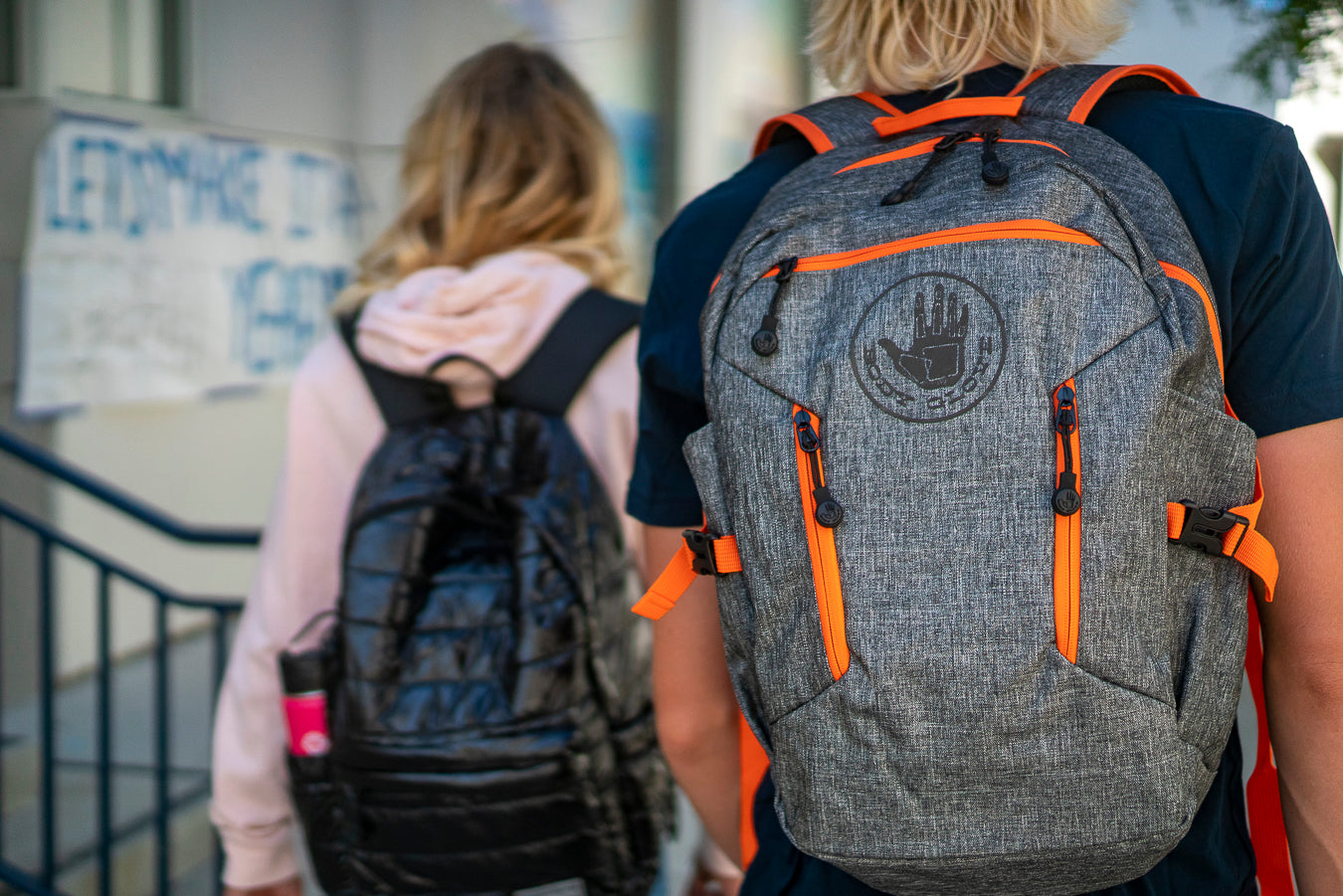 Two people with backpacks walking outdoors, one with a gray and orange backpack.