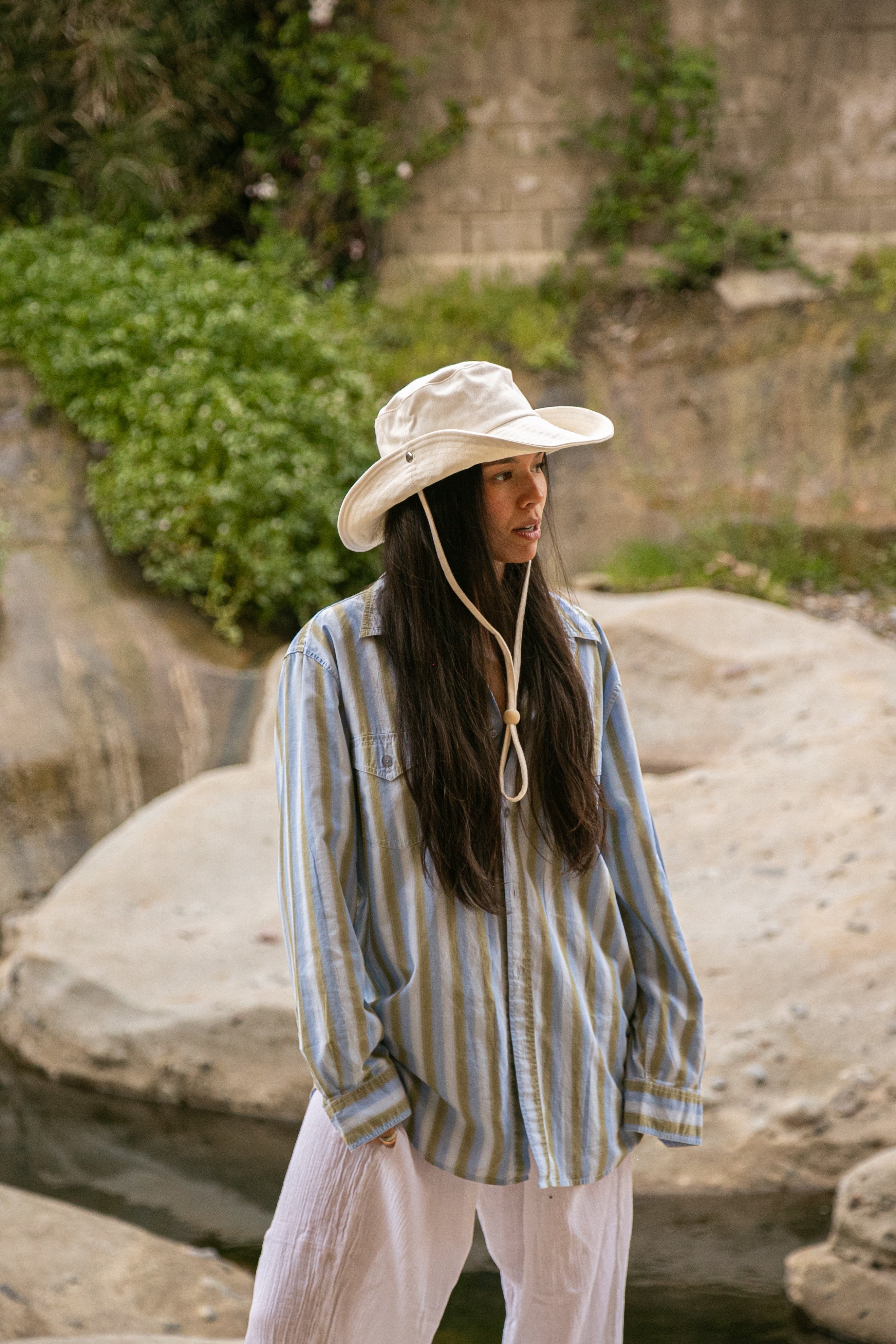 Person wearing a wide-brimmed hat and striped shirt standing near rocks and greenery
