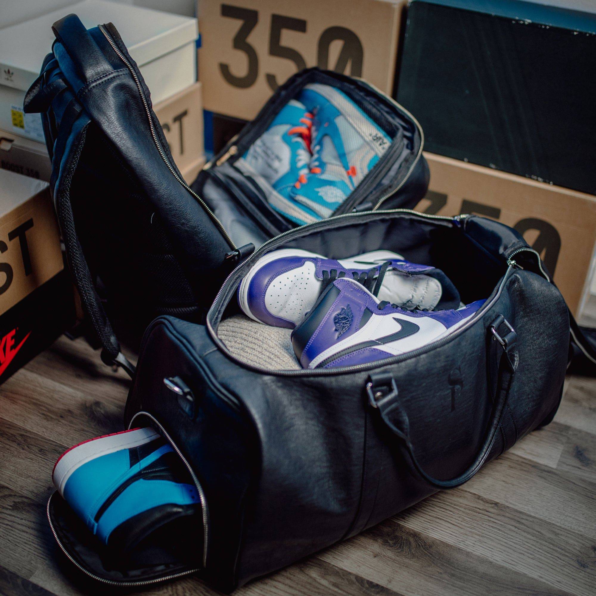 Black duffel bag with blue and white sneakers on a wooden floor with cardboard boxes in the background.