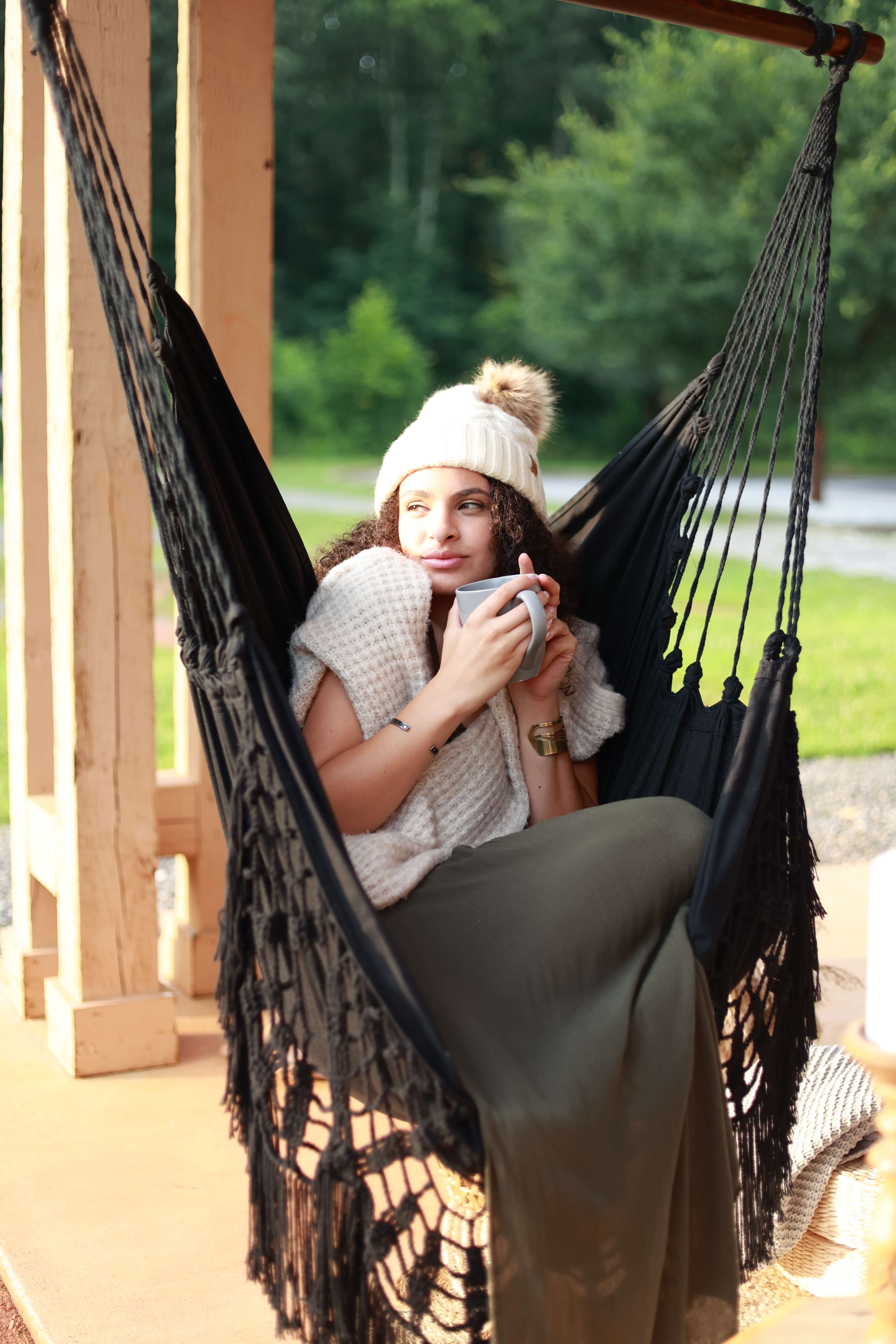 Woman sitting in a black hammock holding a mug, surrounded by greenery.