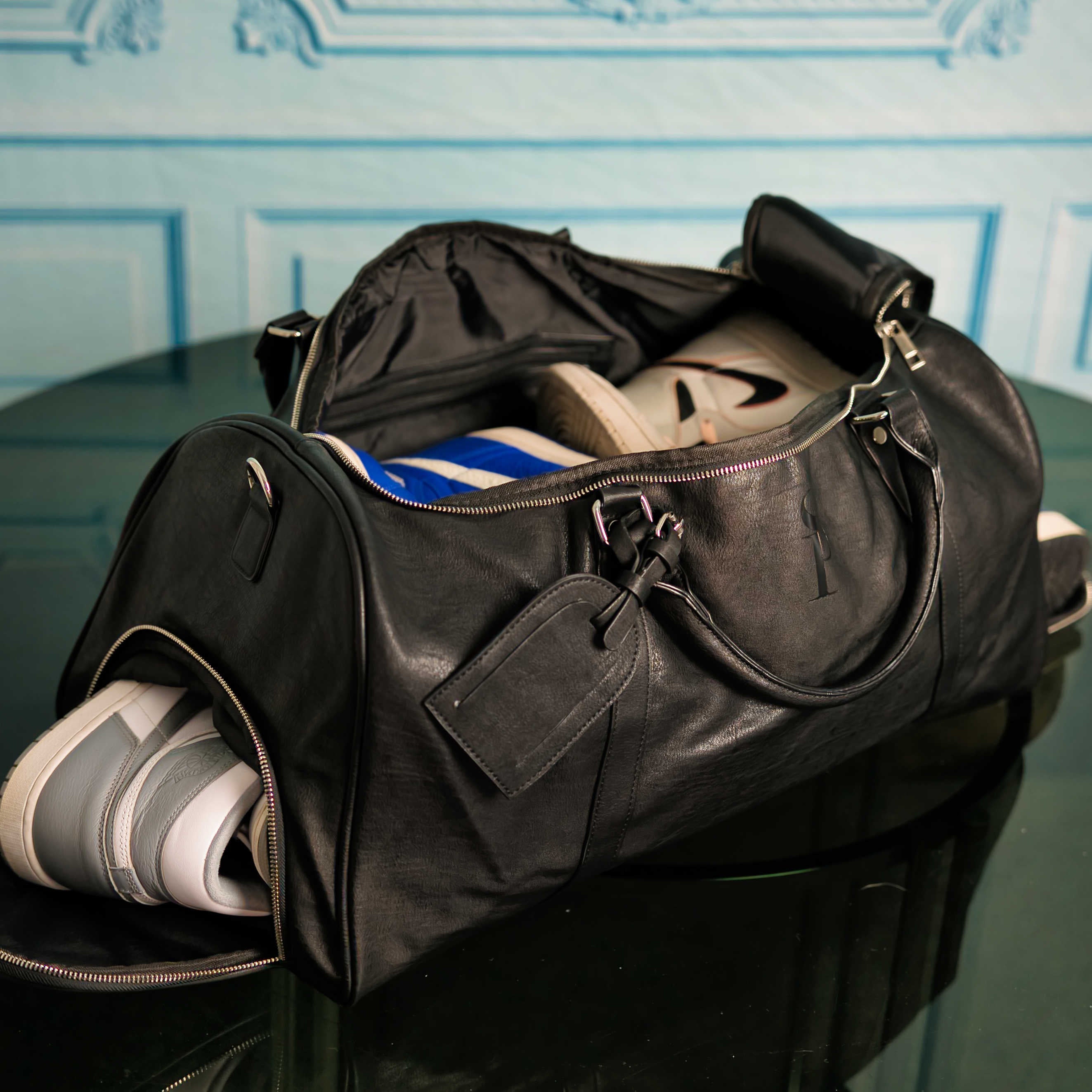 Black leather duffel bag with shoes on a reflective surface against a decorative wall.