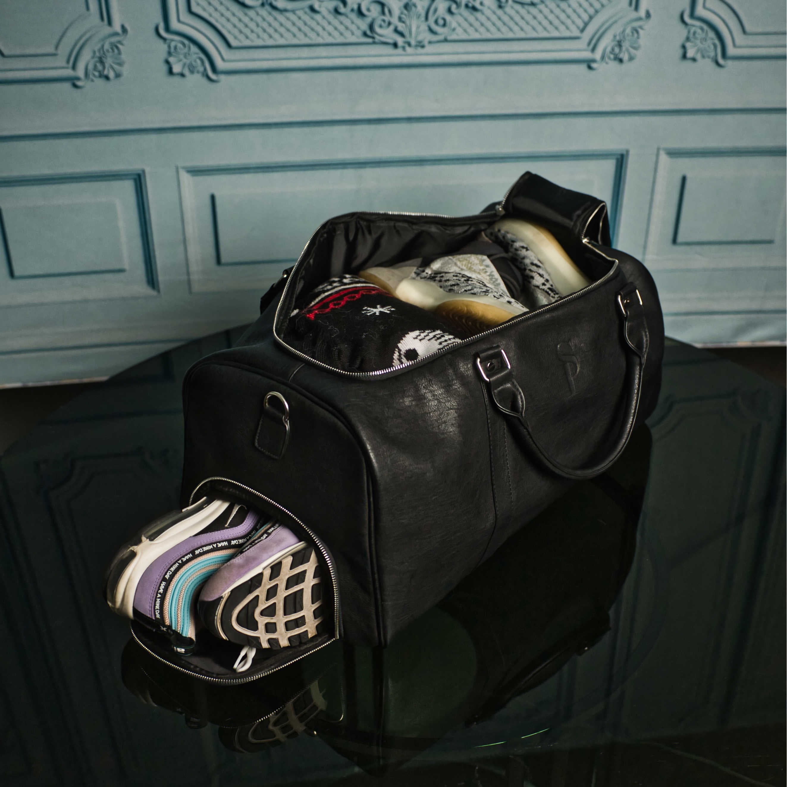 Black duffel bag with colorful shoes on a reflective surface against a decorative wall.