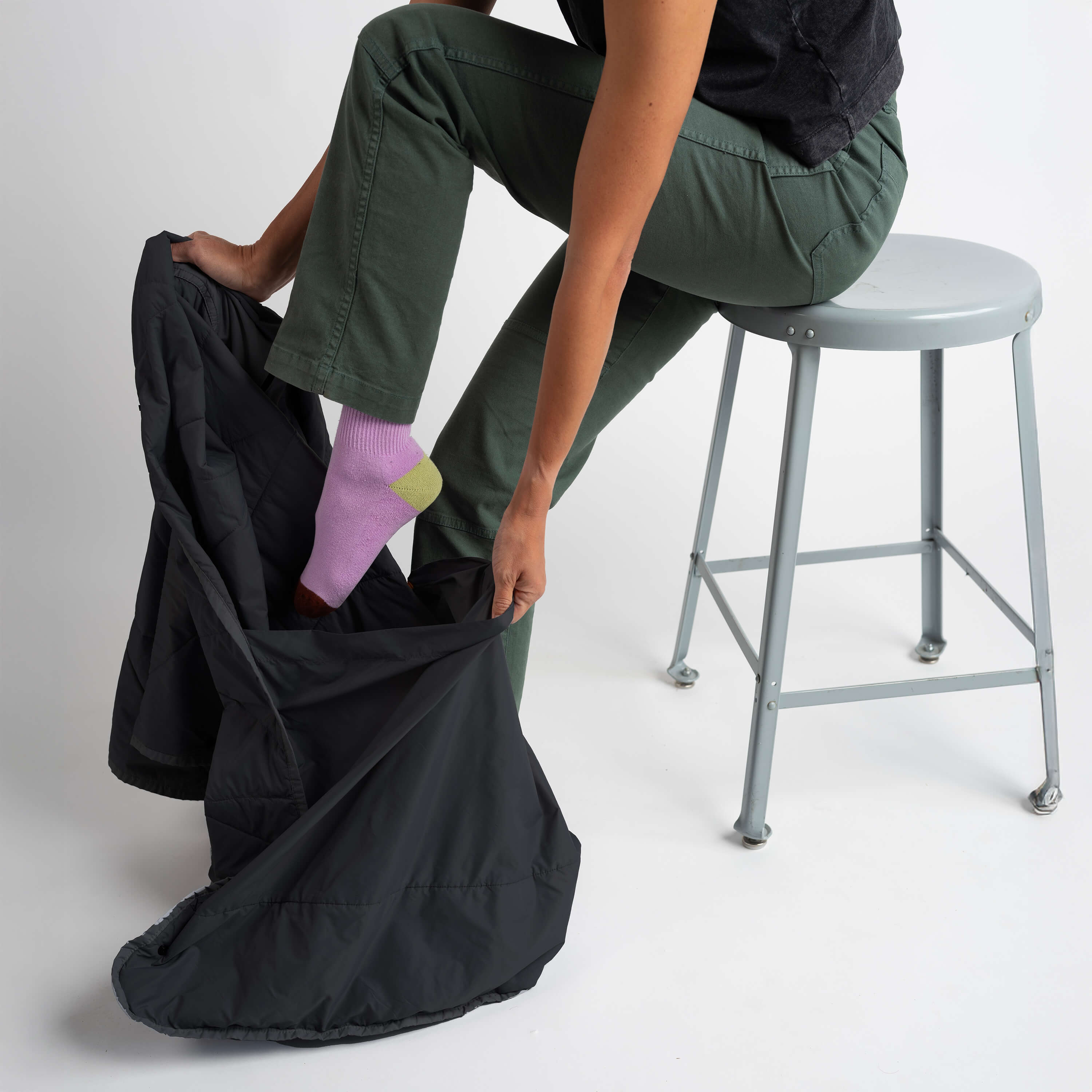 Person sitting on a stool with one leg in a black bag, wearing green pants and pink socks.