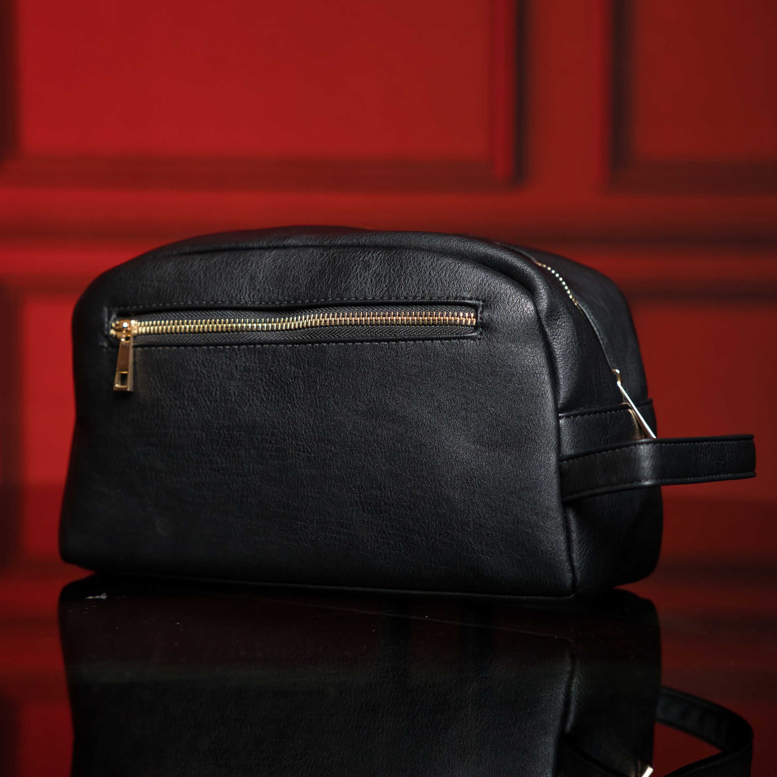 Black leather toiletry bag with gold zipper on a reflective surface with red background