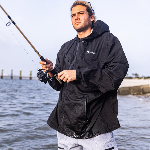Man wearing a black jacket with a brand logo, holding a fishing rod by a body of water.