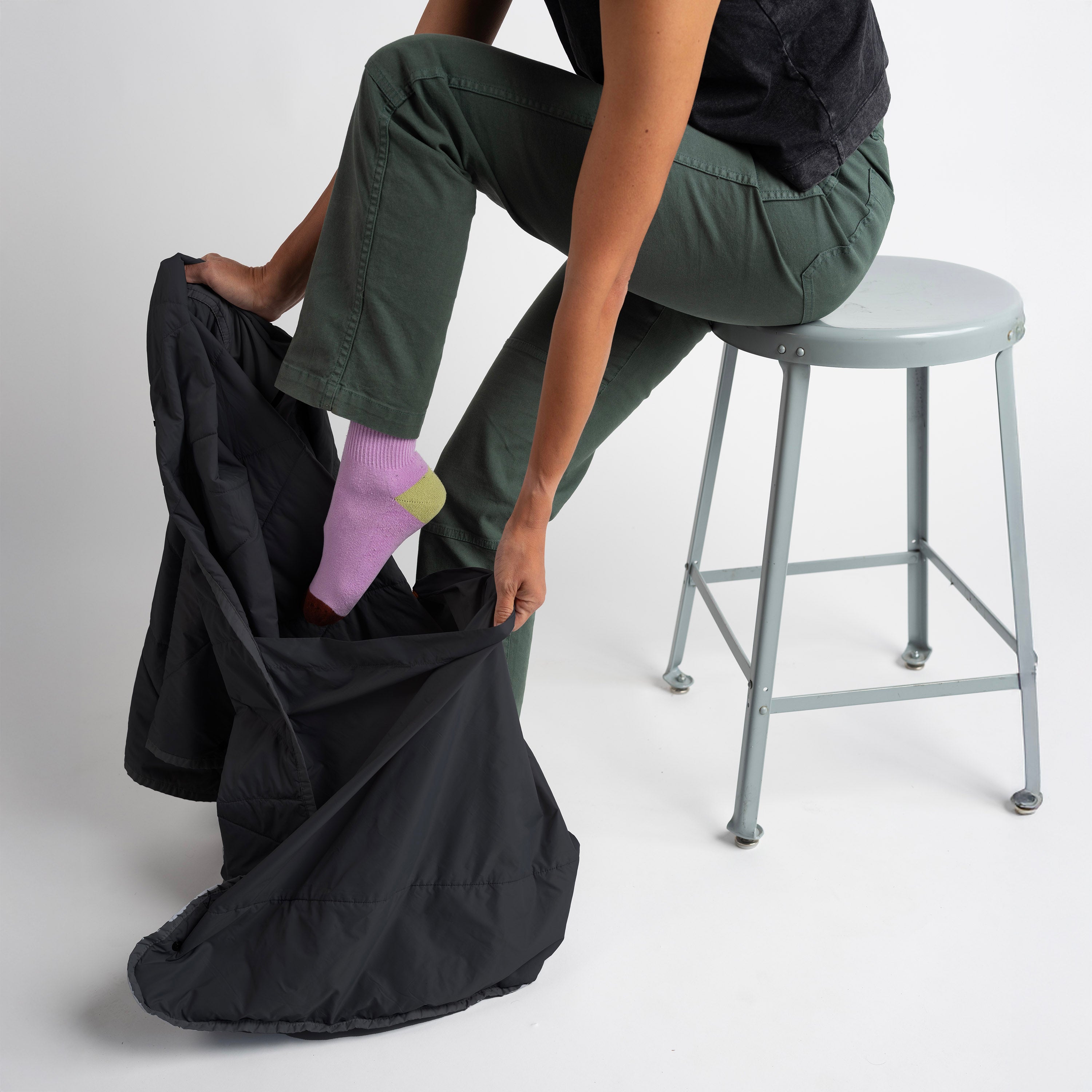 Person sitting on a stool with one foot in a black bag, wearing green pants and pink socks.
