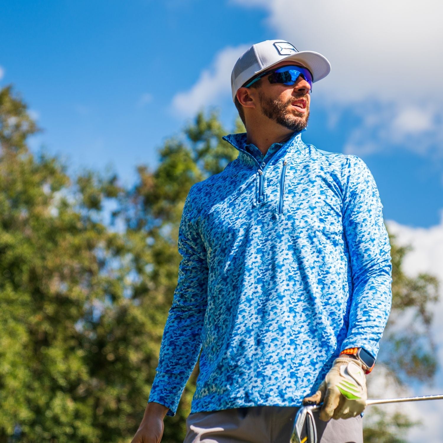 Man in a blue patterned shirt and white cap holding a golf club outdoors.
