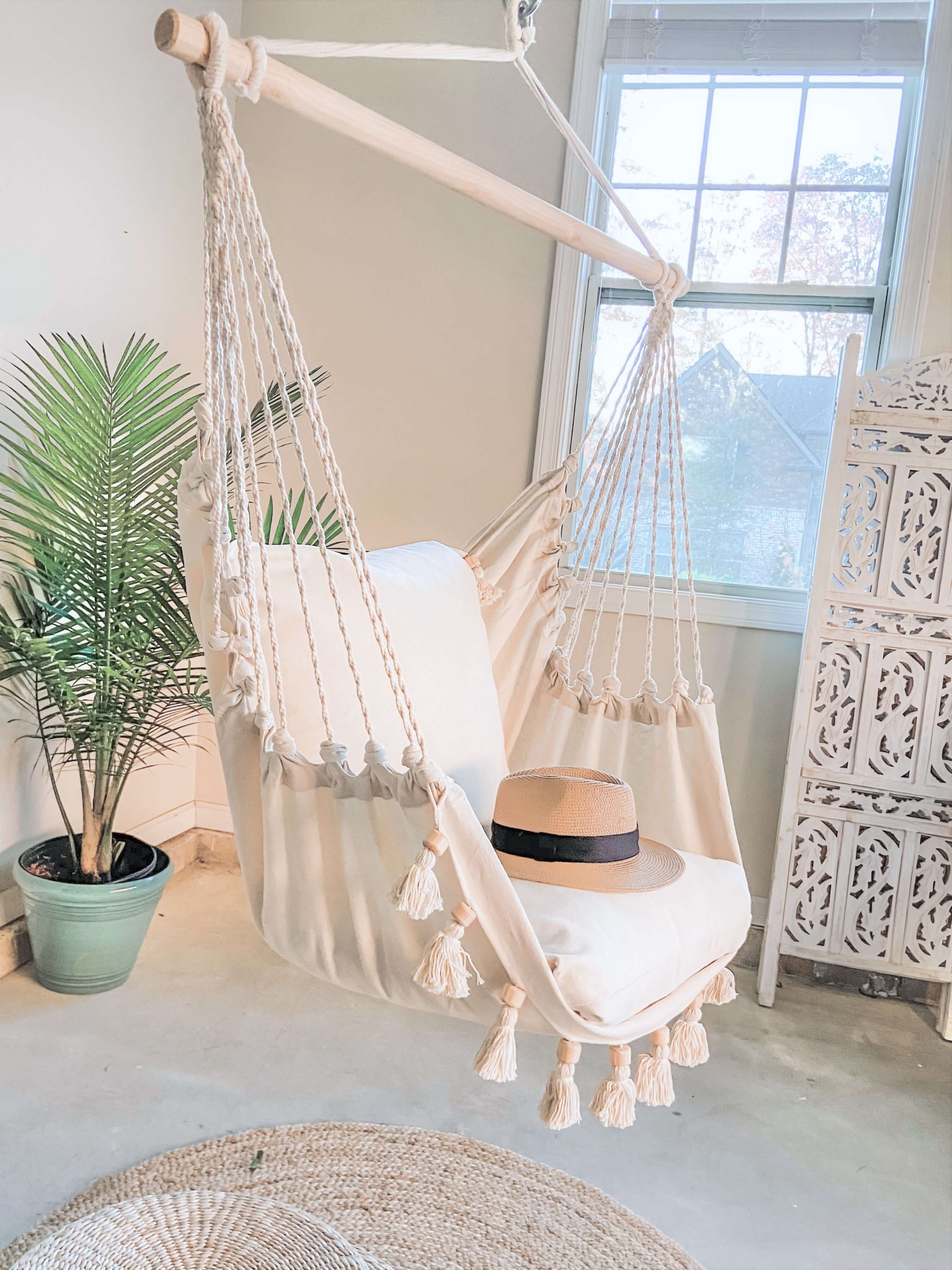 White hammock chair with tassels in a room with a plant and decorative cabinet.