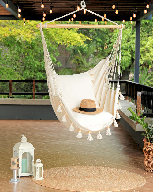 White hammock chair with tassels on a wooden deck with a hat and lanterns.