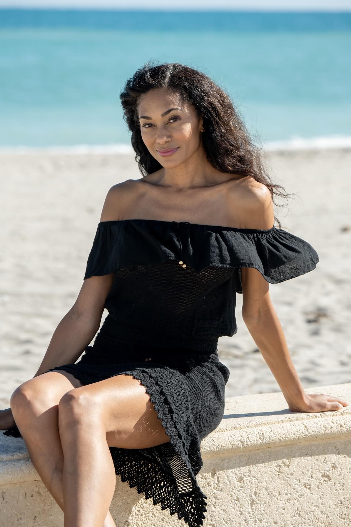 Woman in a black off-shoulder dress sitting on a beach.