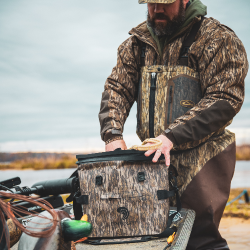 Man in camouflage gear opening a cooler by a body of water.