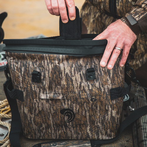 Camouflage cooler bag with a person's hand opening it, set against a blurred natural background.