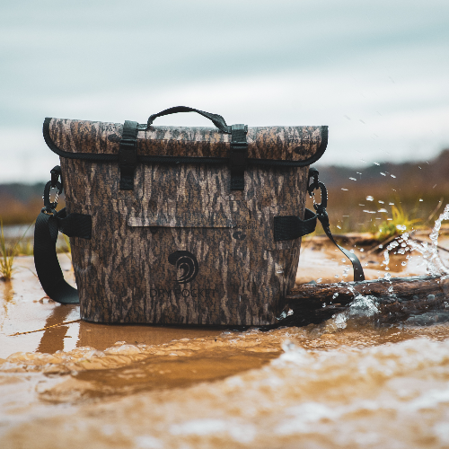 Camouflage-patterned cooler bag in a natural setting with water splashing around it.