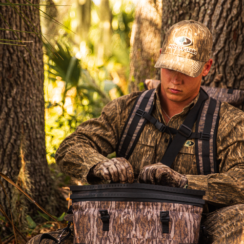 Person in camouflage gear sitting in a forest with a camouflage cooler.